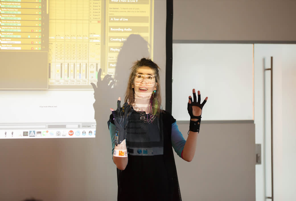 A woman stands in front of a projected computer screen, her face and hands illuminated by the projection. She is smiling, wearing dark clothing and gloves, and gesturing with both hands raised.