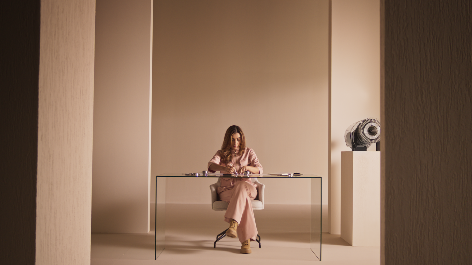 A woman in pink business attire sits at a glass desk in a minimalist beige office, focused on paperwork. A sculptural object is displayed on a pedestal to her right.