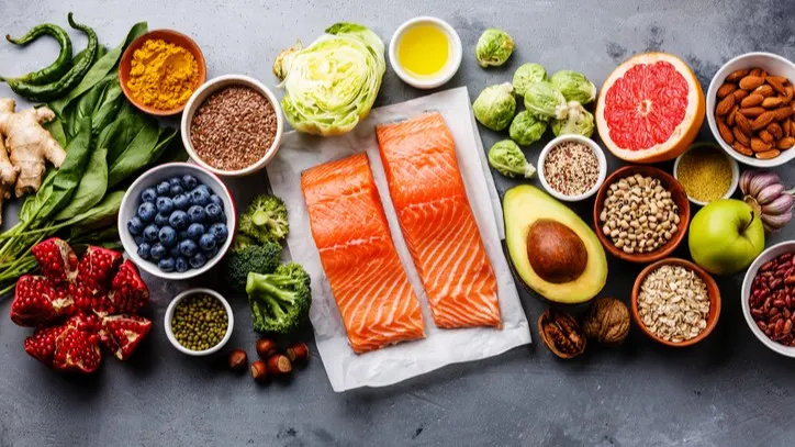 A variety of healthy foods on a gray surface, including salmon fillets, avocado, berries, grapefruit, leafy greens, nuts, seeds, grains, spices, and vegetables arranged in a colorful display.