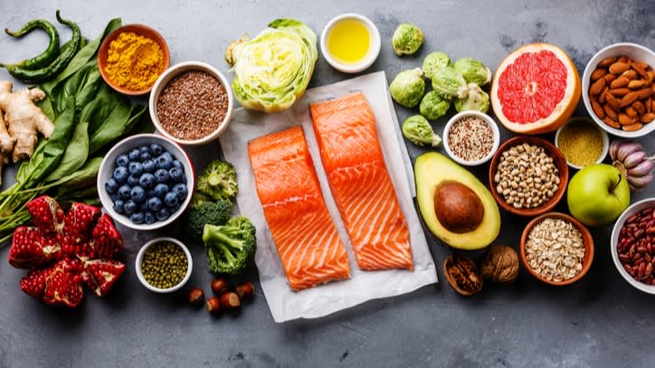 A variety of healthy foods on a gray surface, including salmon fillets, avocado, berries, grapefruit, leafy greens, nuts, seeds, grains, spices, and vegetables arranged in a colorful display.