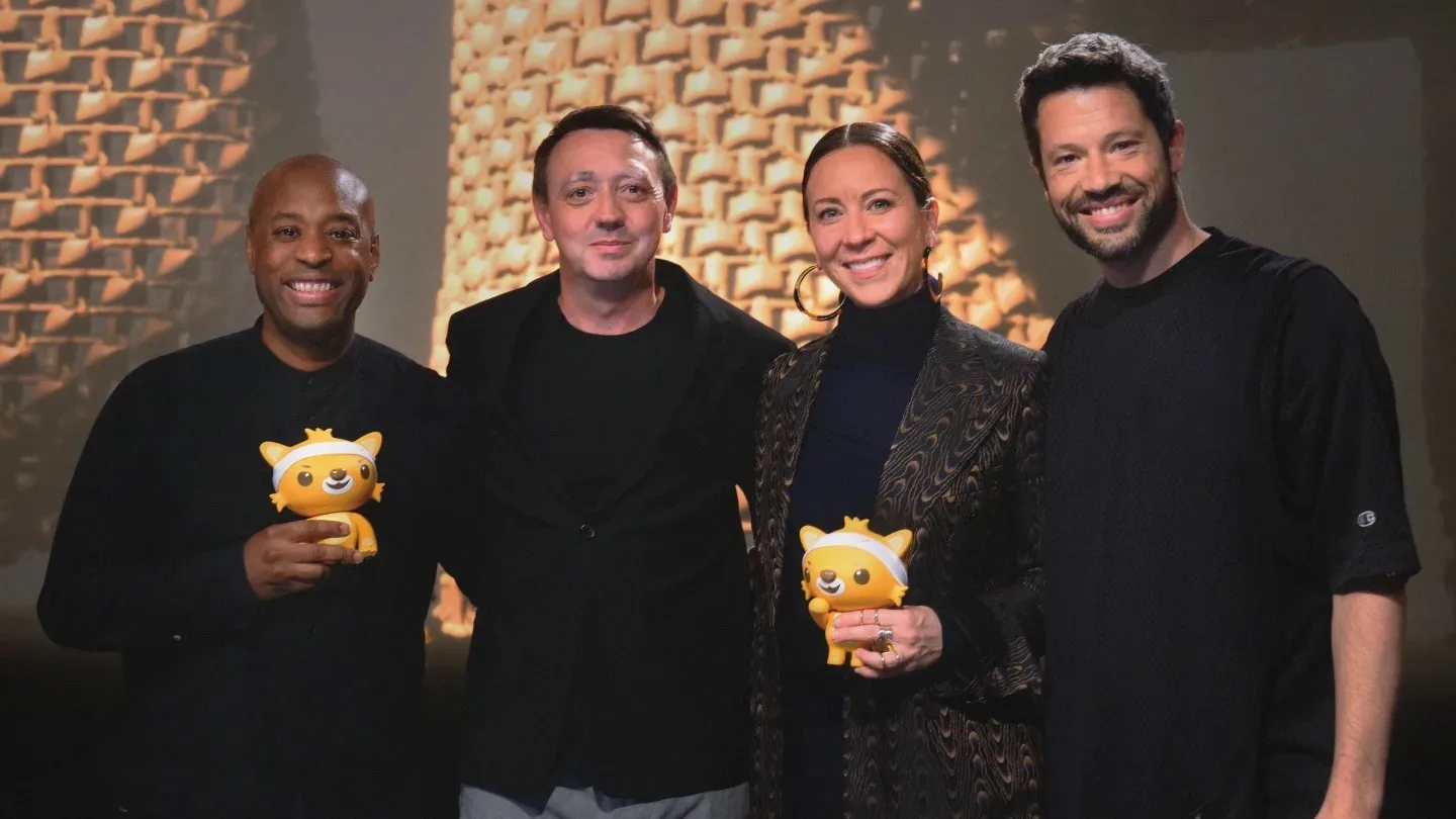 Four people stand together smiling. Two of them hold small, yellow, animal-shaped trophies. The background features a pattern of repeating geometric shapes.