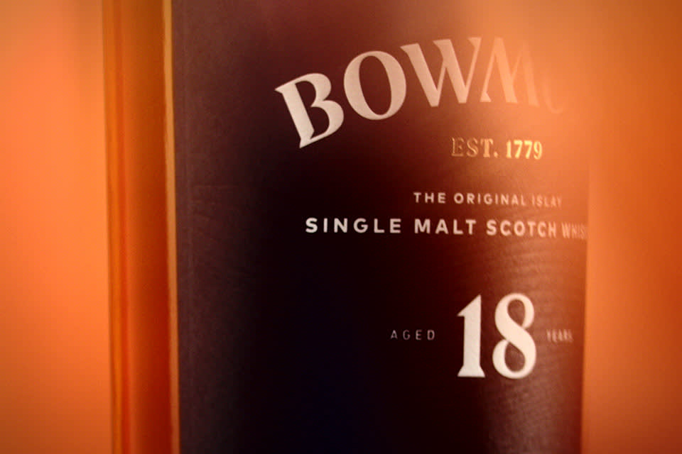 Close-up of a Bowmore Single Malt Scotch Whisky bottle label, highlighting the text BOWMORE, EST. 1779, Single Malt Scotch, and Aged 18 Years against a warm, orange-toned background.