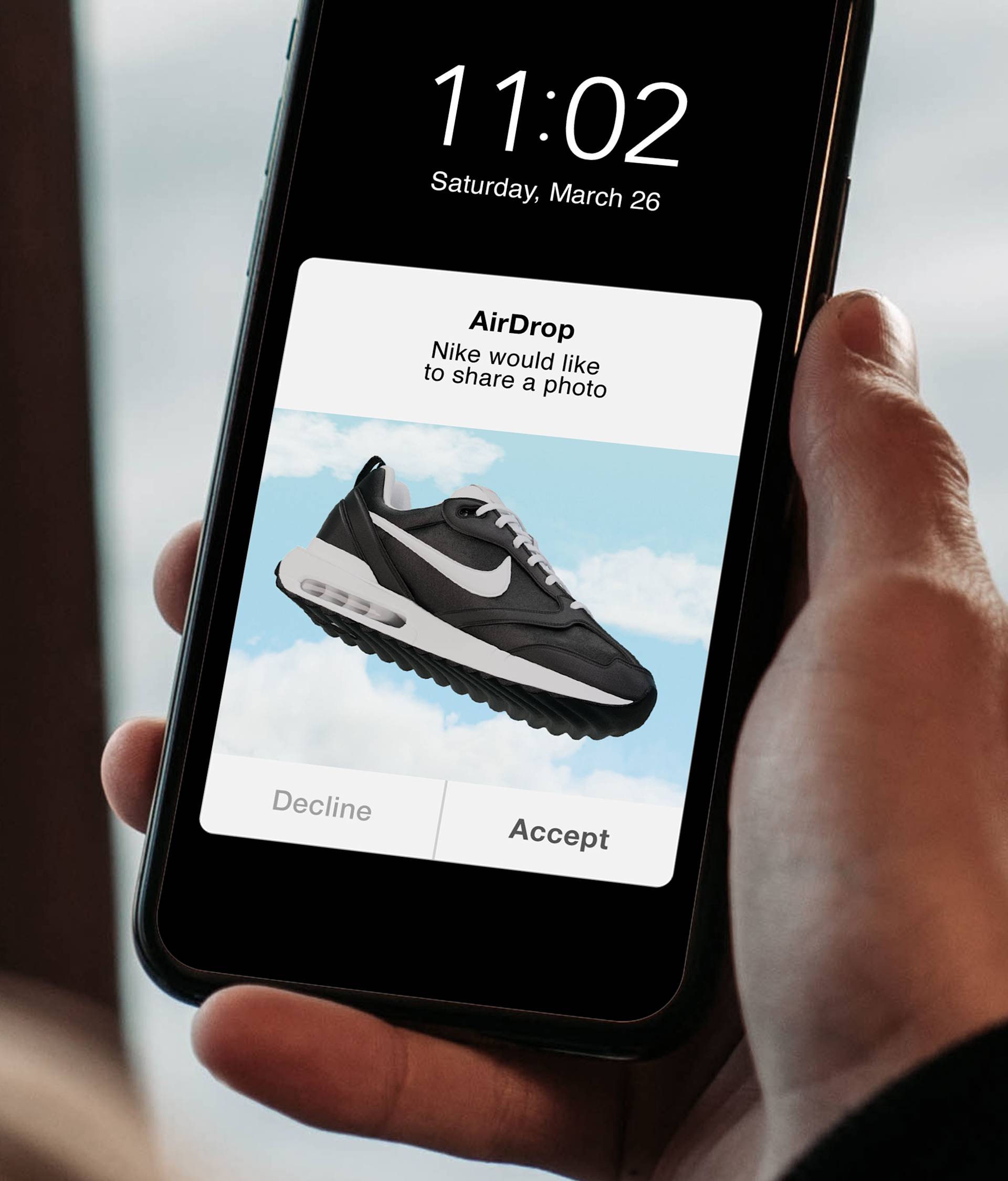A hand holding a smartphone showing an AirDrop request; the request displays a photo of a black and white Nike sneaker against a sky background, with Decline and Accept buttons visible below.