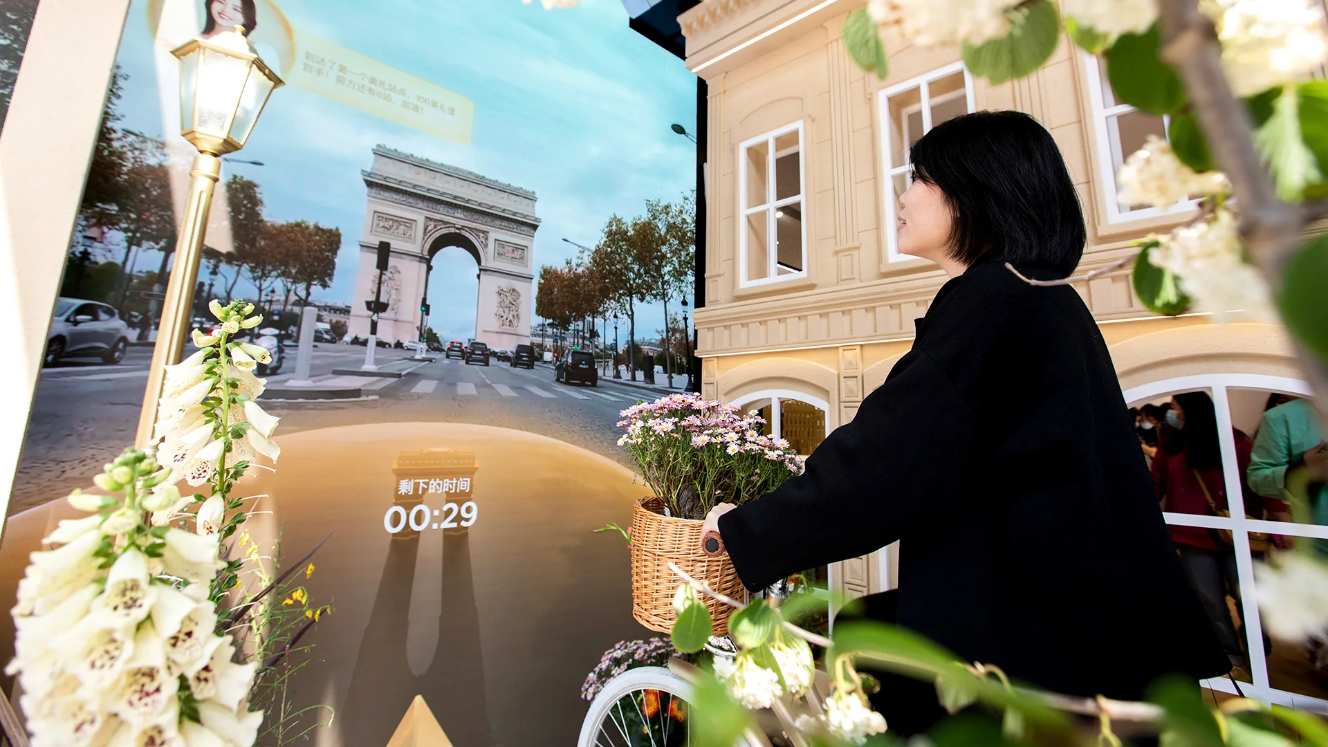 A woman with a bicycle basket of flowers faces a large screen displaying the Arc de Triomphe and a Parisian street, surrounded by floral decorations and buildings resembling a European setting.