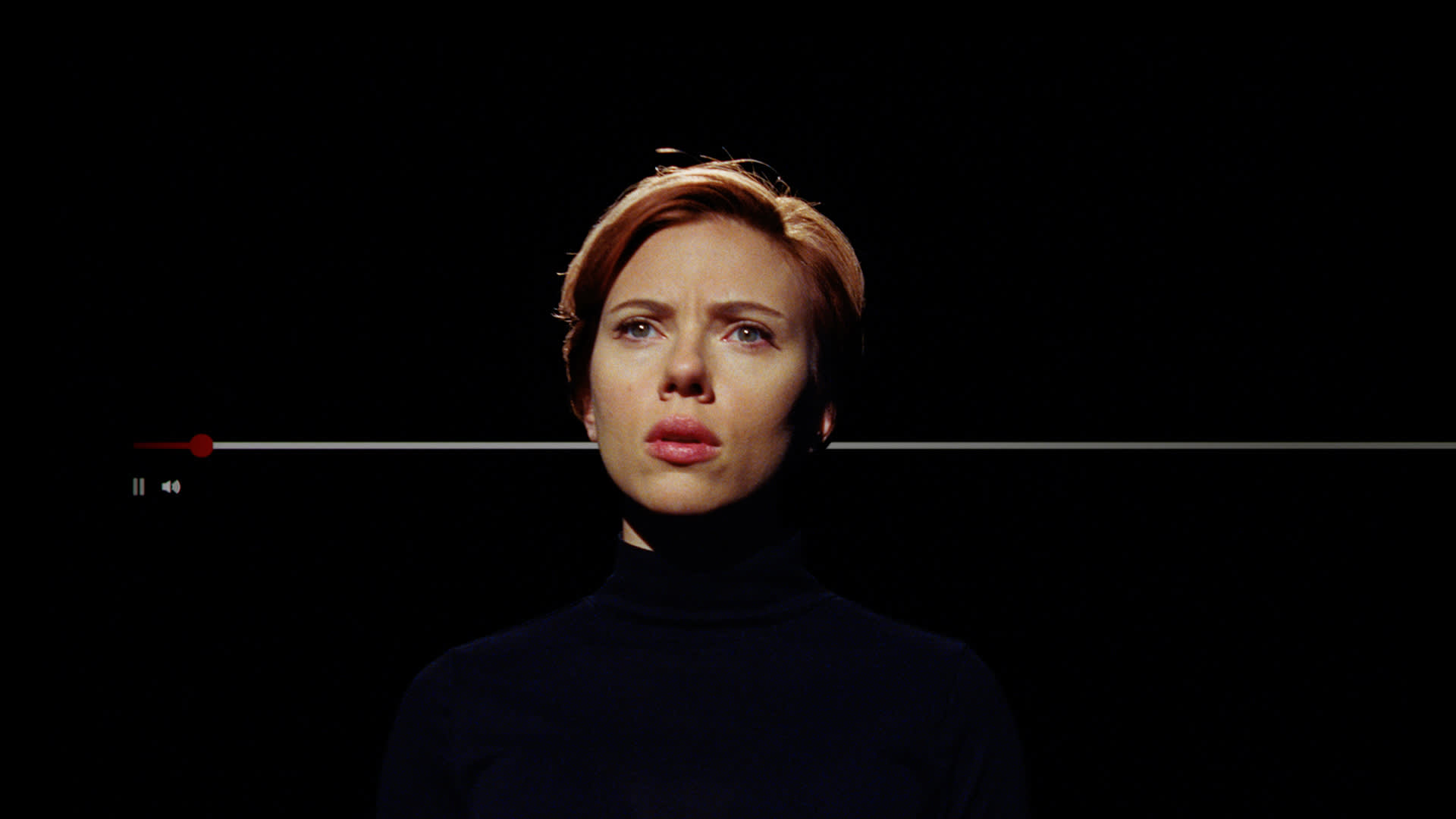 A woman with short, reddish hair wearing a black turtleneck looks serious under dramatic lighting, with a video pause bar overlaid across the image against a dark background.