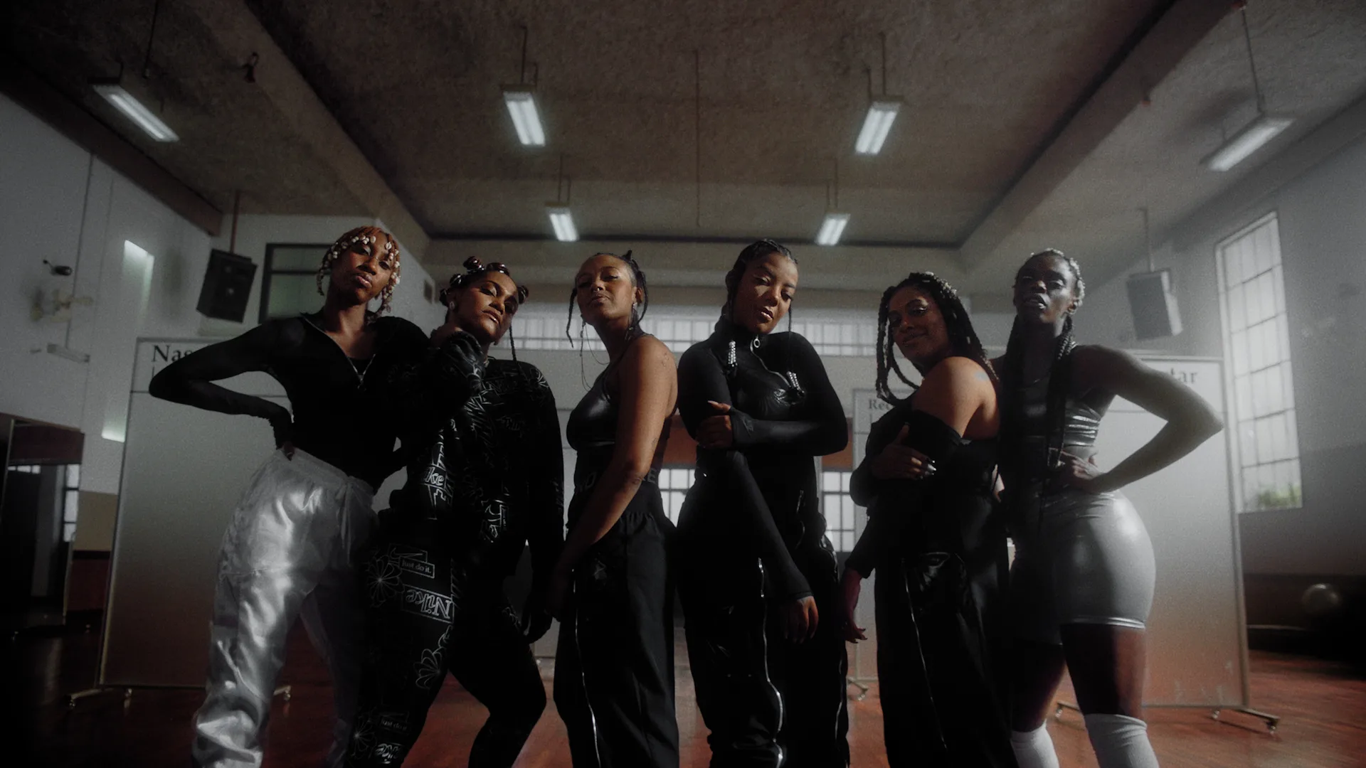 Six women stand confidently in a row inside a spacious, well-lit room with high ceilings and large windows, wearing stylish black and white outfits with bold, expressive poses.