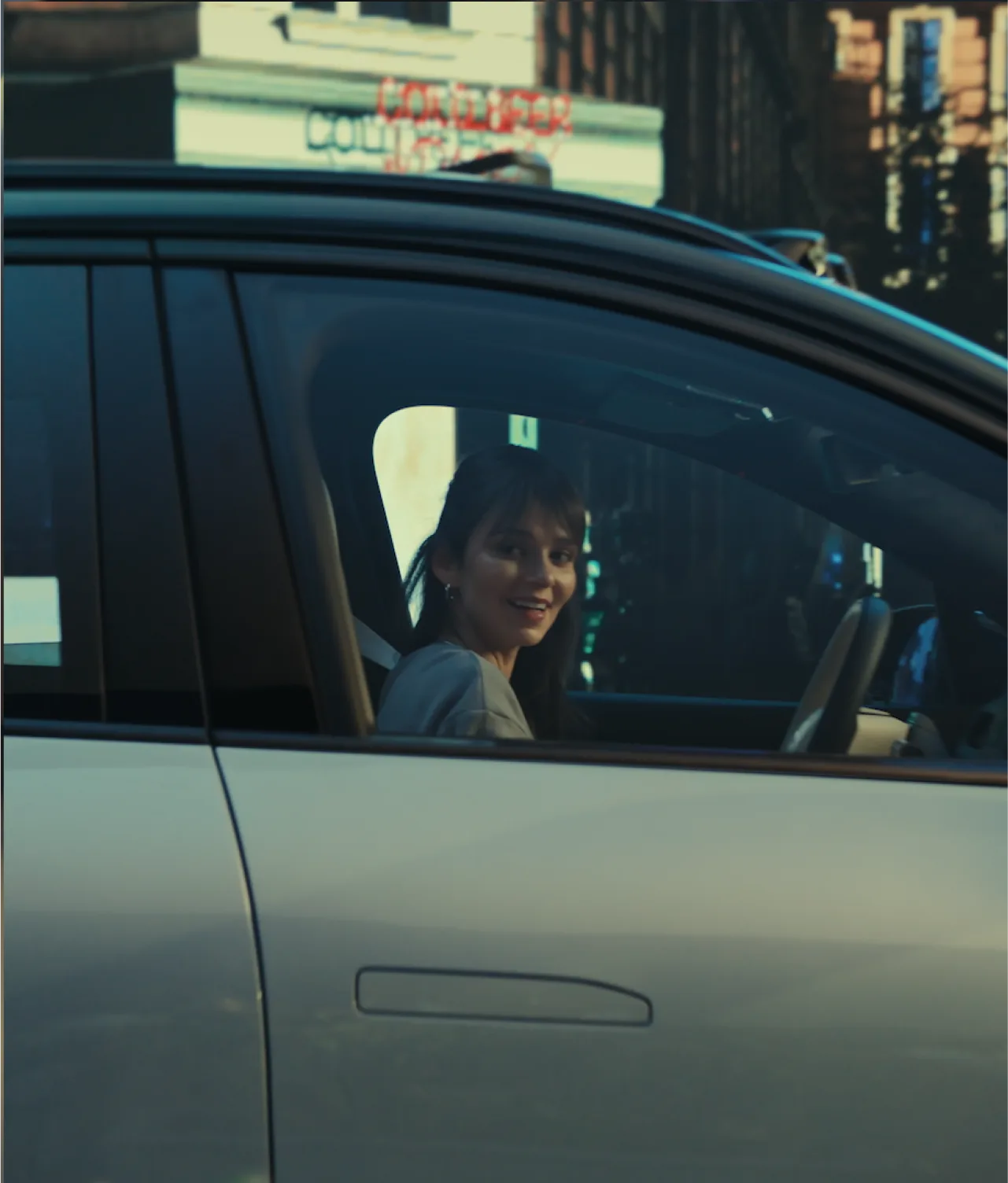 A woman with dark hair smiles while sitting in the drivers seat of a car, looking out the open window. The background shows a blurred building and neon signage.