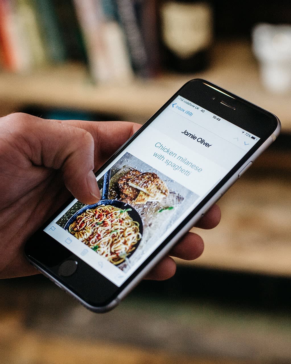 A person holds a smartphone displaying a recipe page titled “Chicken Milanese with Spaghetti,” featuring a photo of cooked spaghetti and chicken in a pan. Books are blurred in the background.