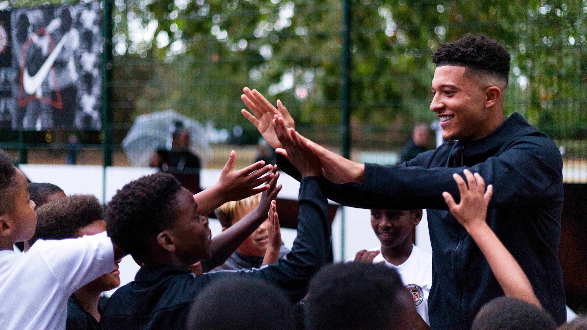 A smiling young man high-fives a group of enthusiastic children outdoors, with a wire fence and trees in the background. The scene is lively and joyful, suggesting a community or sports event.