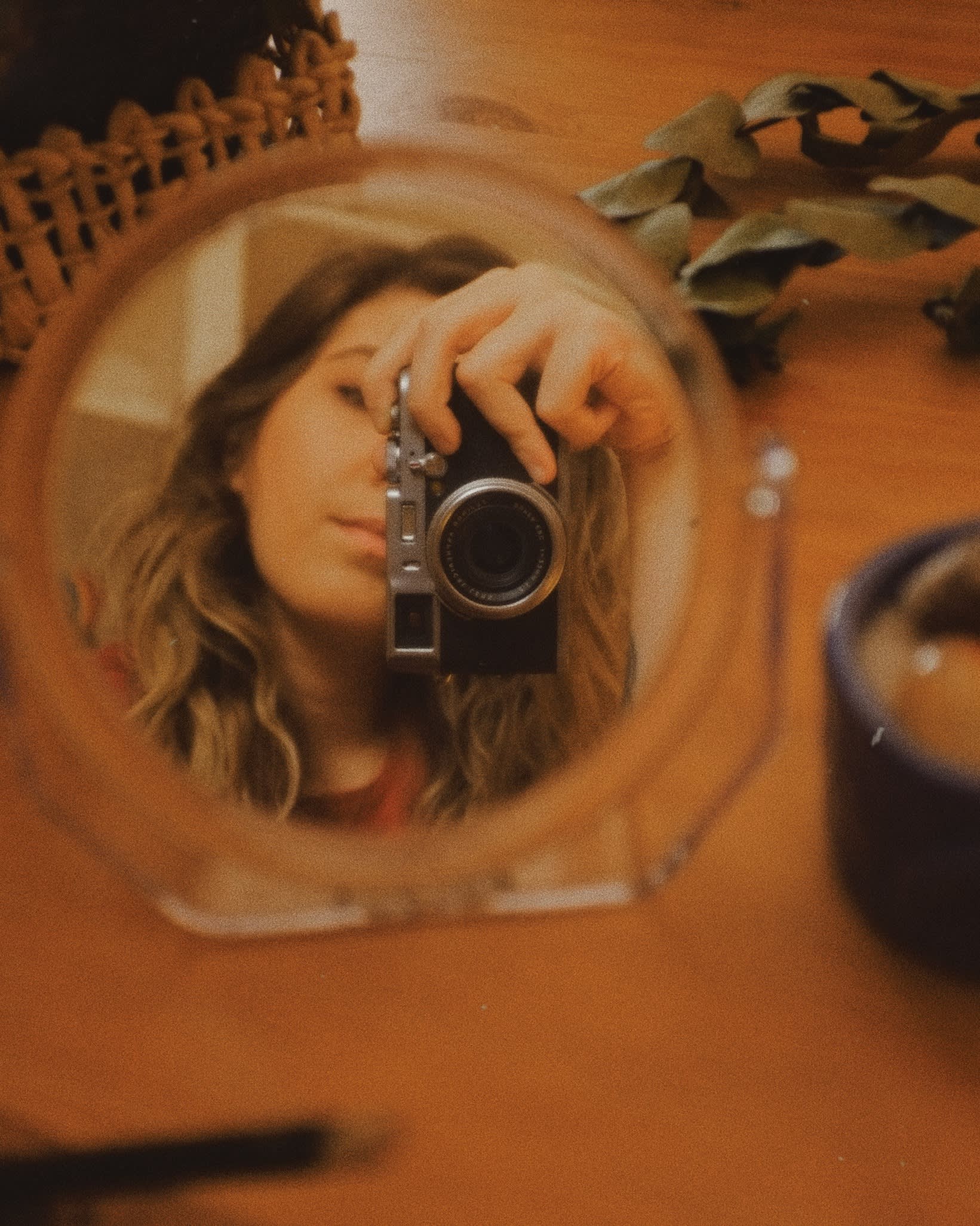 A person with wavy hair takes a photo of themselves using a camera, their reflection visible in a small round mirror on a wooden surface with scattered items around.