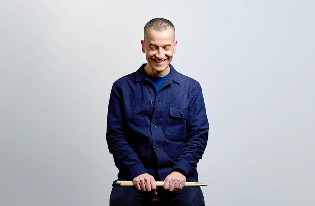 A smiling man with short hair wearing a navy blue jacket sits against a plain light background, looking down and holding drumsticks in both hands.