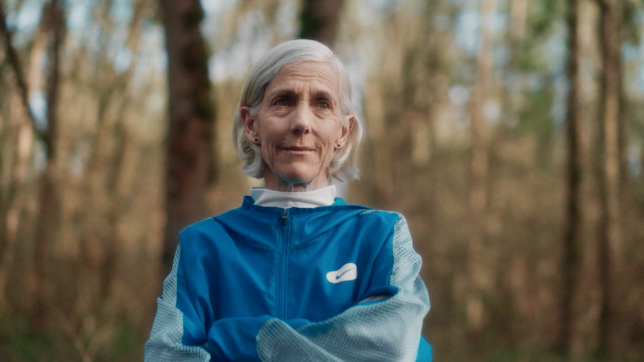 An older woman with gray hair, wearing a blue athletic jacket, stands outdoors with arms crossed, looking confident. Blurred trees and sunlight form the background.