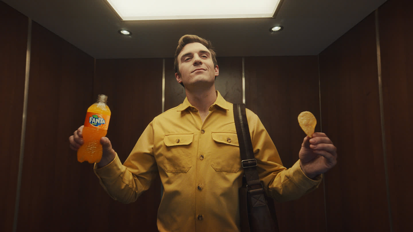 A man in a yellow shirt stands in an elevator holding a bottle of orange Fanta in one hand and a potato chip in the other, looking confident under bright overhead lighting.