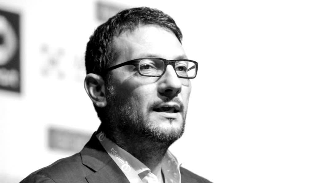A man with short dark hair, glasses, and a beard, dressed in a suit, is speaking or presenting on stage. The background is blurred with indistinct logos and shapes. The image is in black and white.