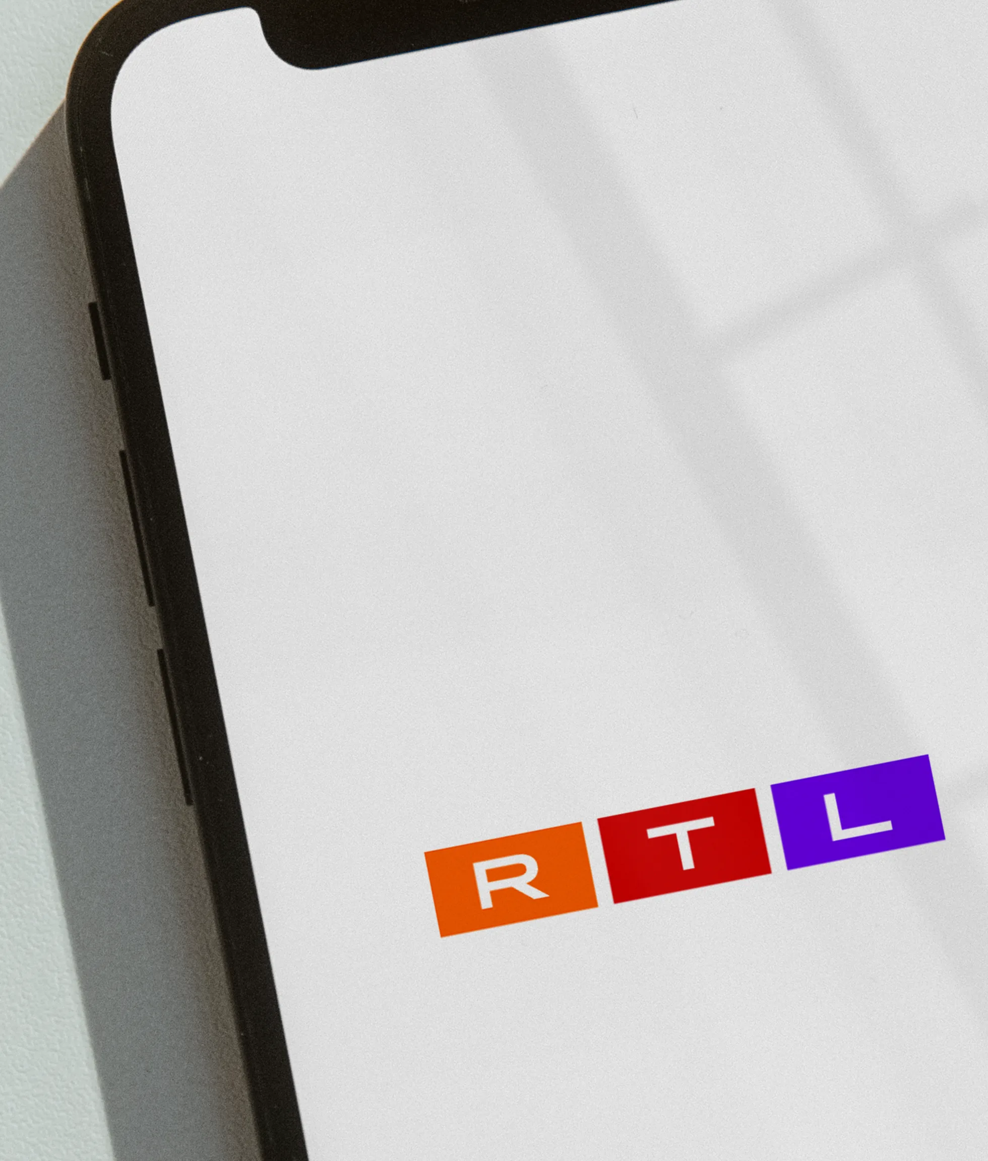 A smartphone screen displays the RTL logo, with the letters R, T, and L in white on orange, red, and purple squares, respectively. The phone is angled, showing part of its black frame.