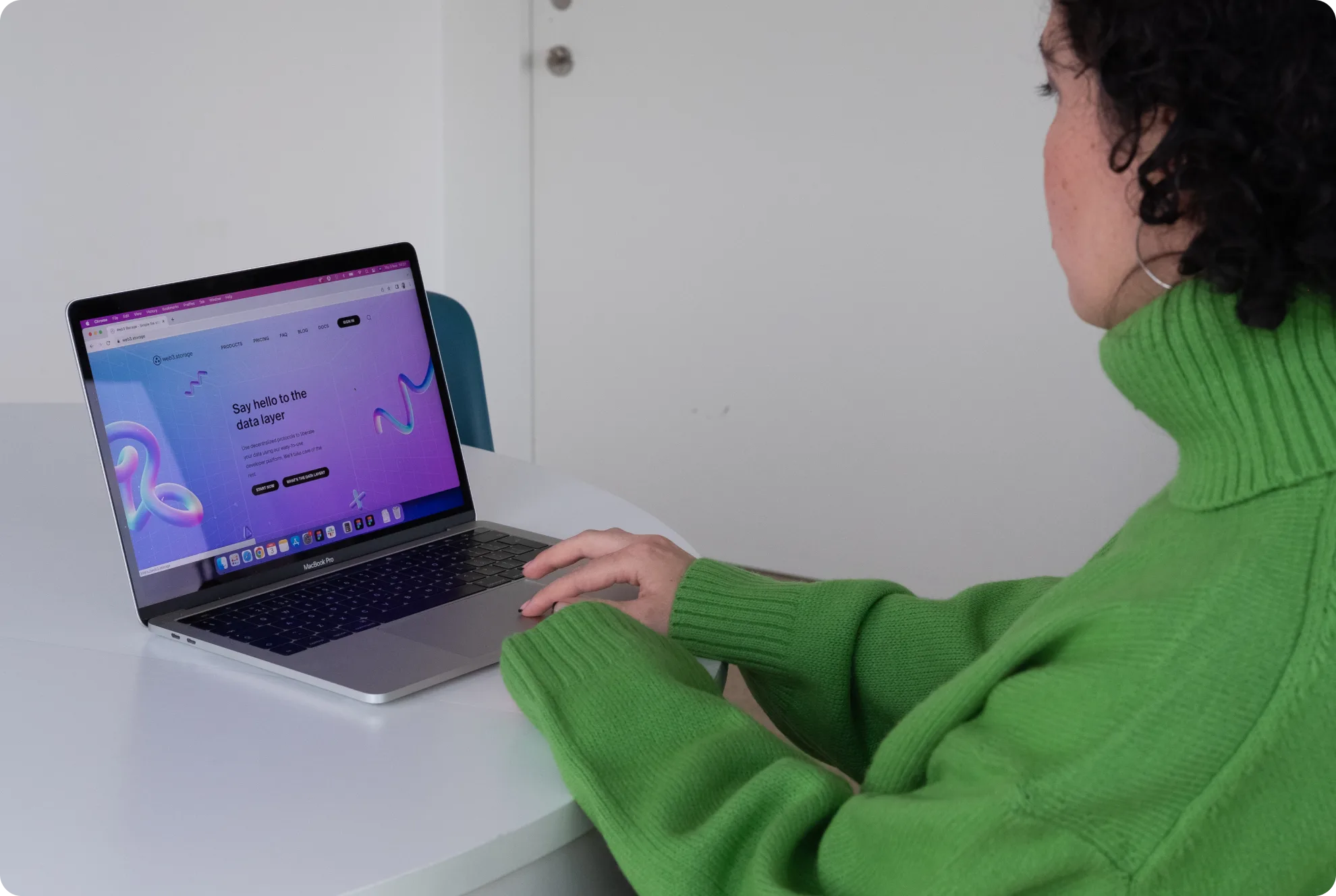 A person wearing a green sweater is sitting at a table, using a laptop. The laptop screen displays a colorful website with text and abstract shapes. The persons face is partially visible.