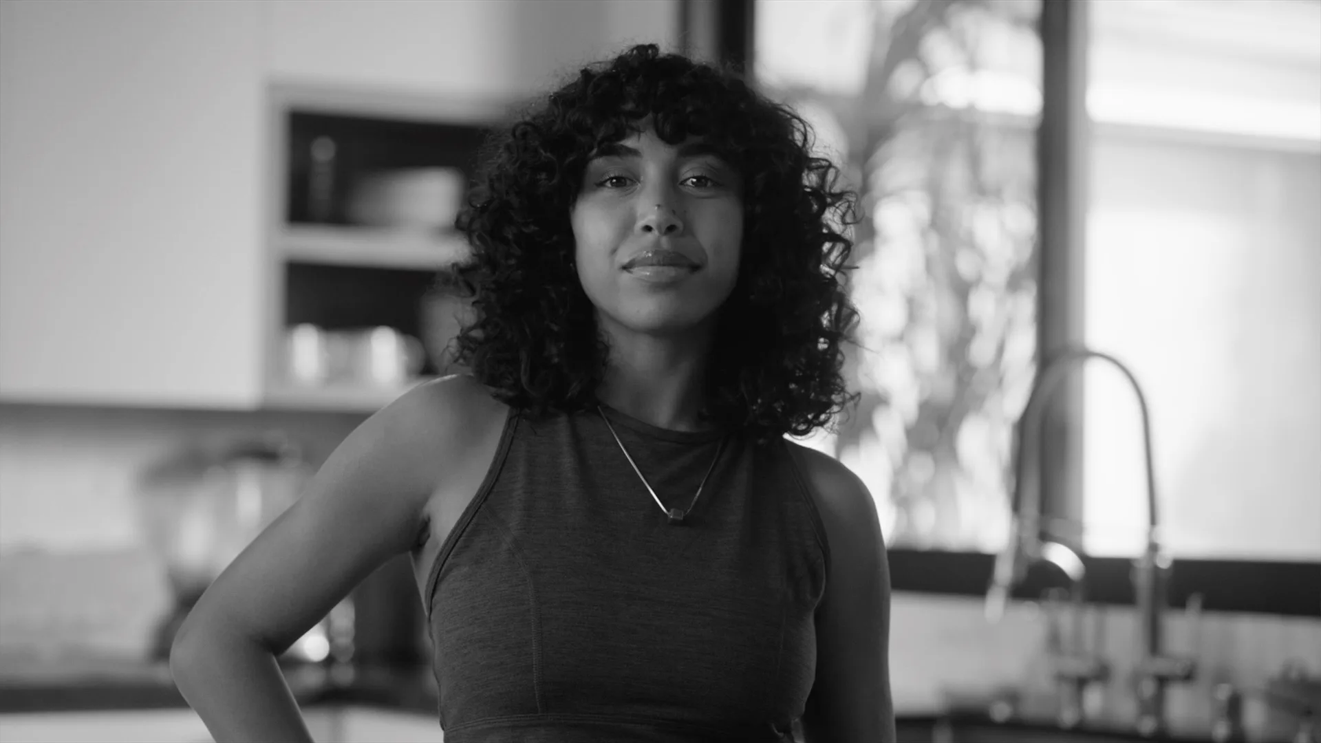 A woman with curly hair wearing a sleeveless top stands confidently in a modern kitchen, looking at the camera. The image is in black and white.