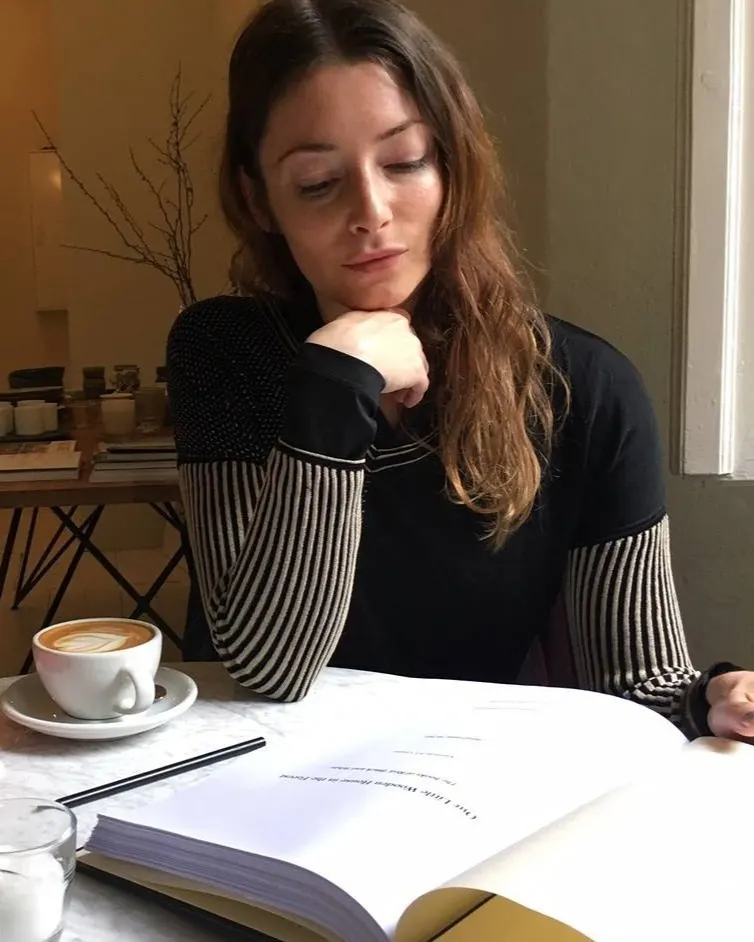 A woman with long brown hair sits at a table by a window, reading a book with her chin resting on her hand. A cup of coffee and a small glass of water are on the table beside her.
