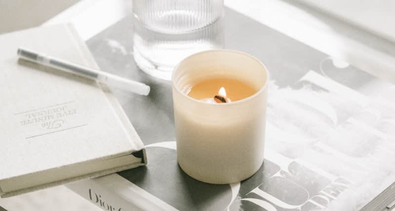 A lit white candle sits on top of a magazine next to a closed journal with a pen and a glass of water, creating a calm and cozy atmosphere.