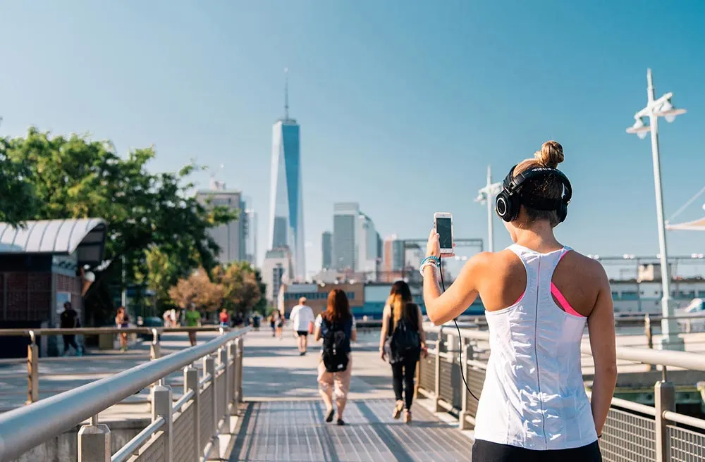 A woman wearing headphones and athletic clothes holds up her phone while walking on a city pathway, with skyscrapers and the One World Trade Center visible in the background under a clear blue sky.