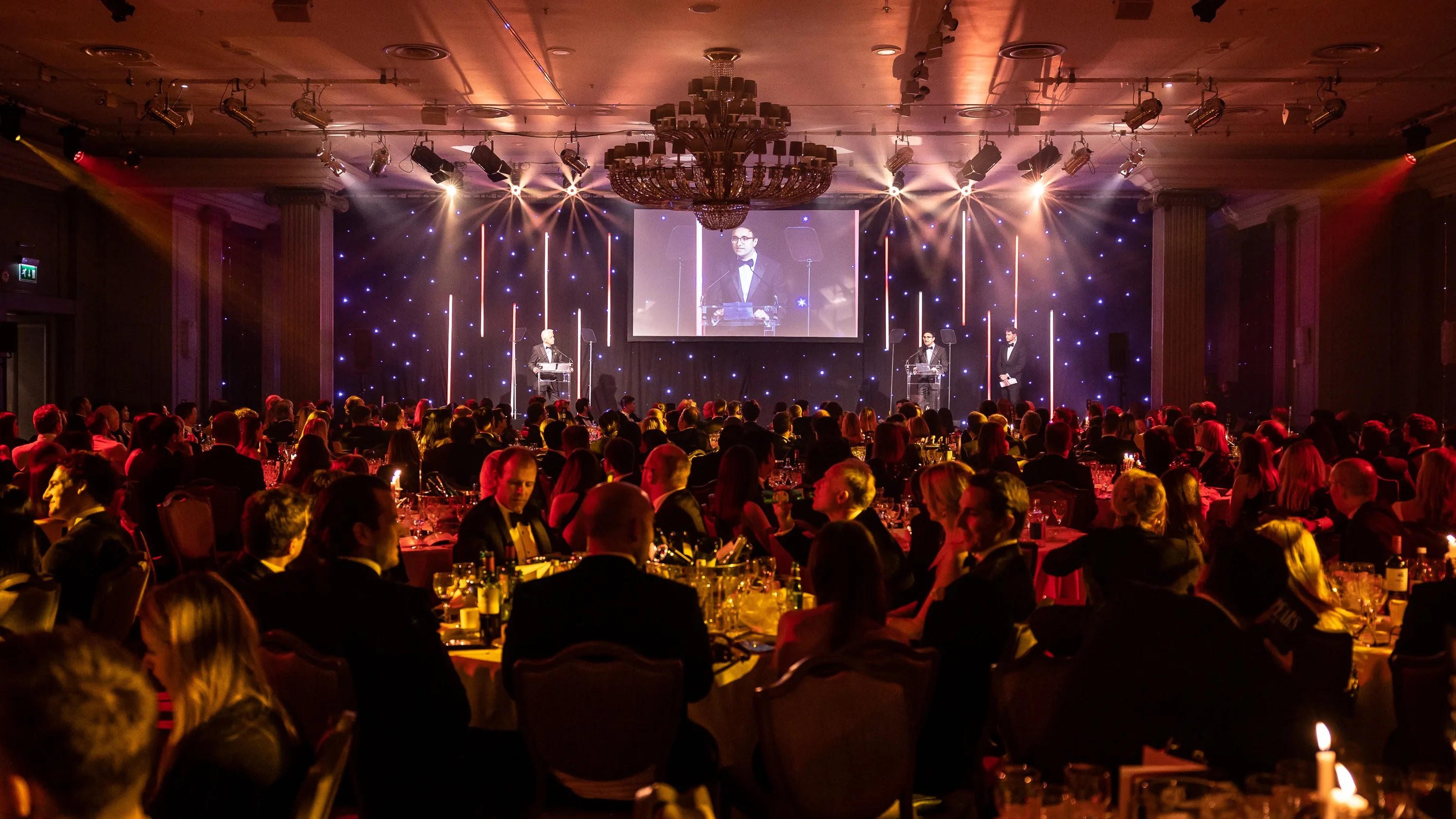A formal banquet with many people seated at round tables, faces lit by warm lighting. A speaker stands at a podium on stage, with their image projected on a large screen behind them. The room glows with elegant, festive lighting.