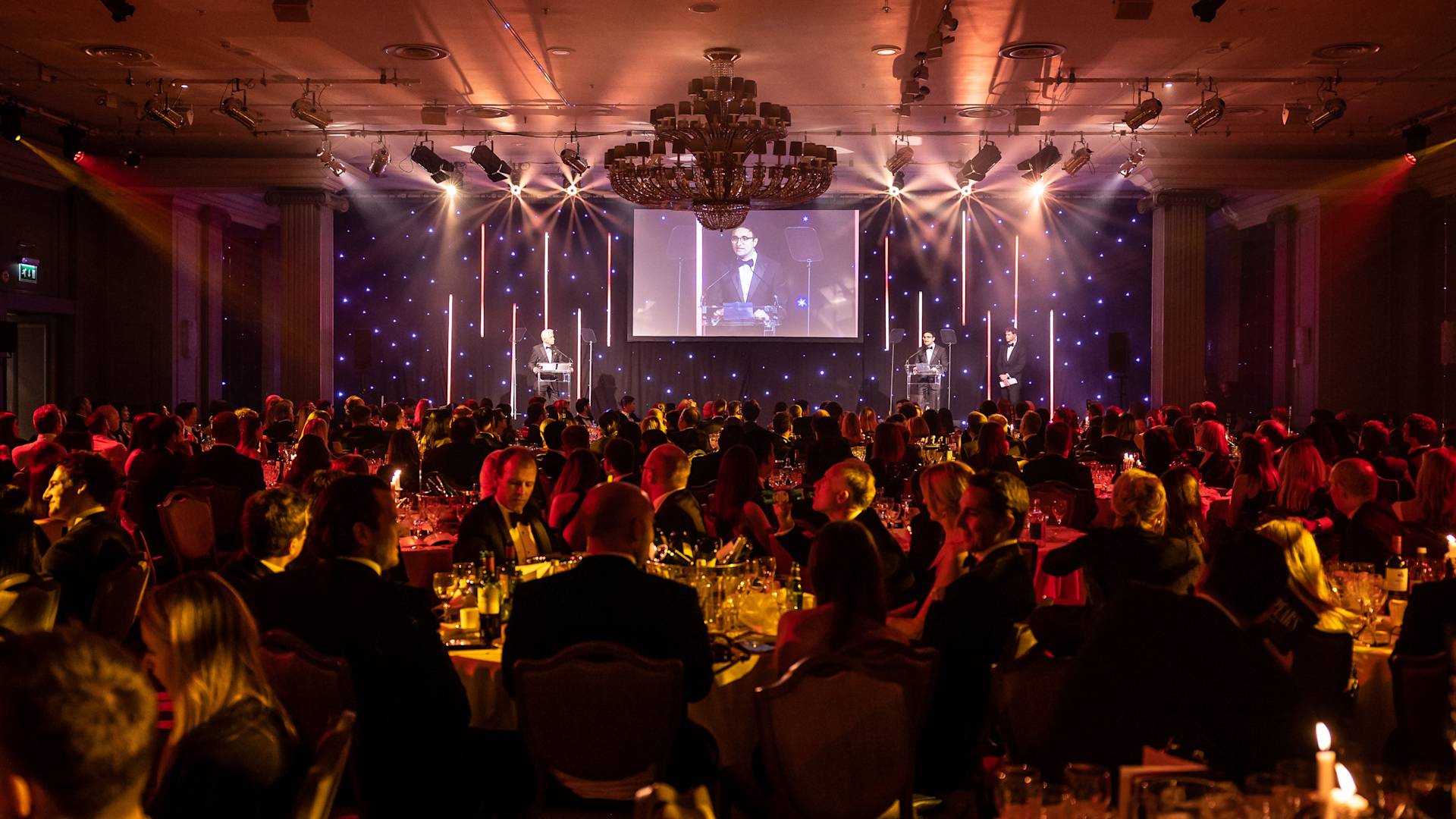 A formal banquet with many people seated at round tables, faces lit by warm lighting. A speaker stands at a podium on stage, with their image projected on a large screen behind them. The room glows with elegant, festive lighting.