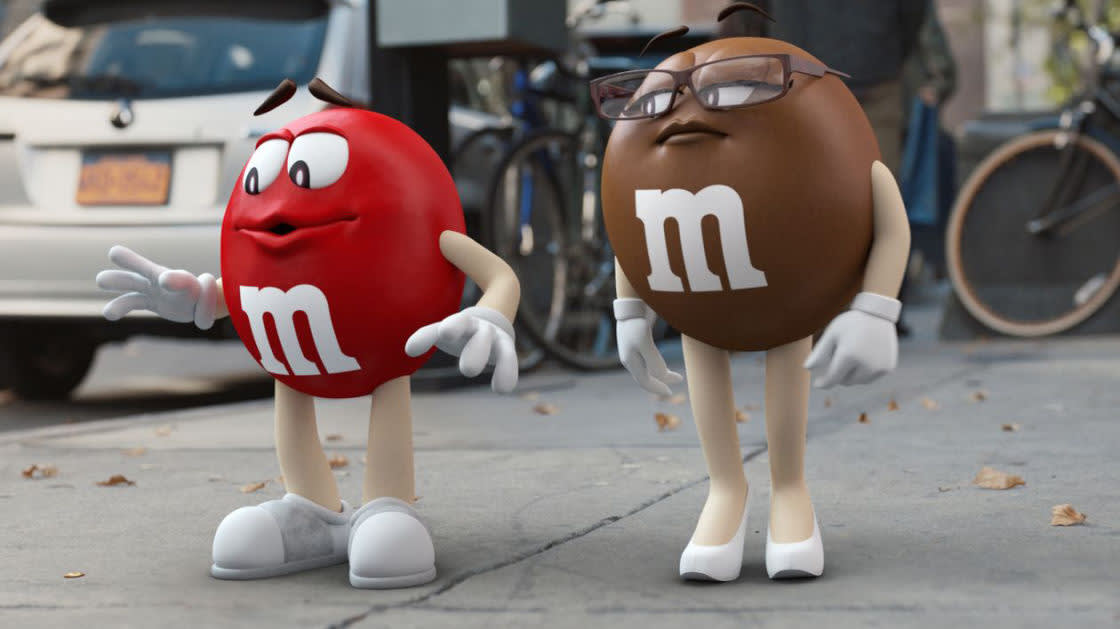 Animated red and brown M&M characters stand on a city sidewalk. The red M&M is gesturing with open arms, while the brown M&M, wearing glasses, stands with hands on hips. Bicycles and cars are visible in the background.