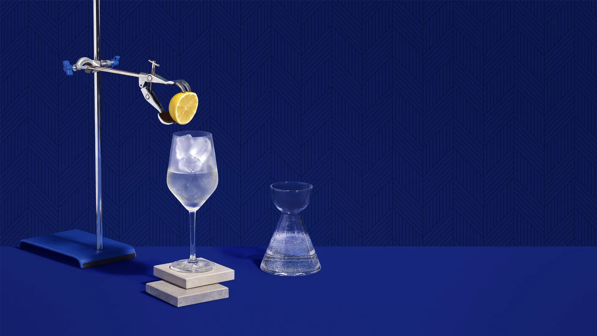A lemon slice held by a lab clamp above a glass of ice water, with a small carafe and stacked coasters, all set on a bright blue surface against a patterned blue background.