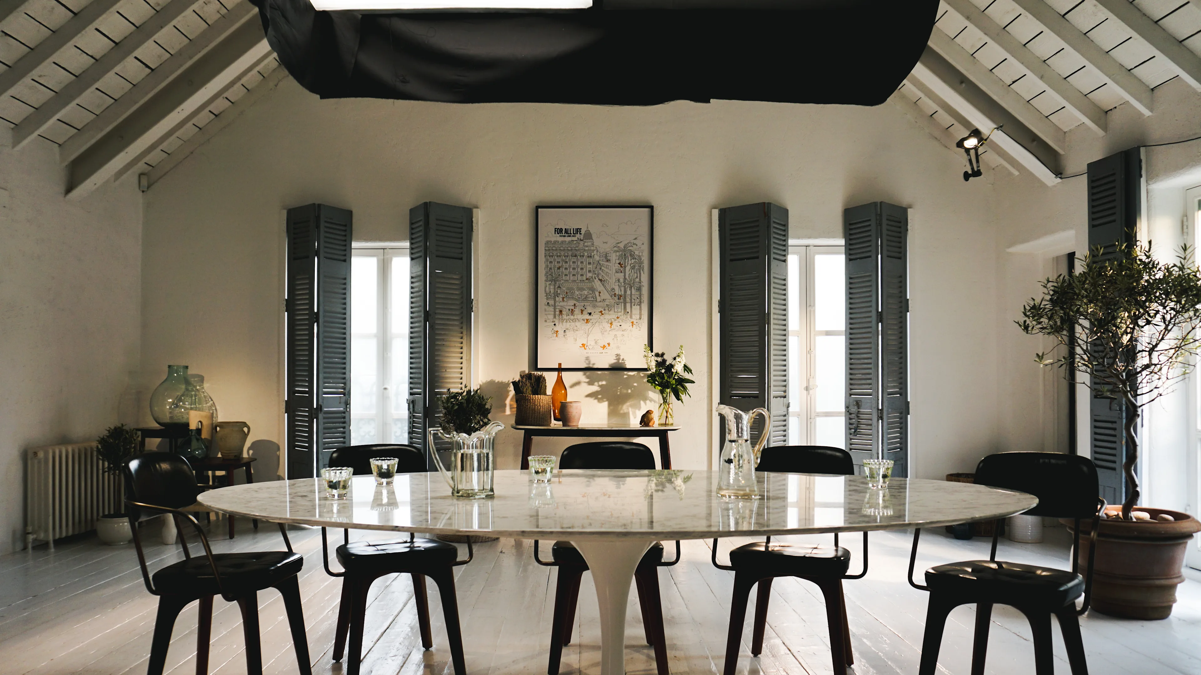 A modern dining room with a large oval marble table, black chairs, glassware, and plants. The space features high ceilings, gray shutters on windows, and a framed artwork on the back wall.