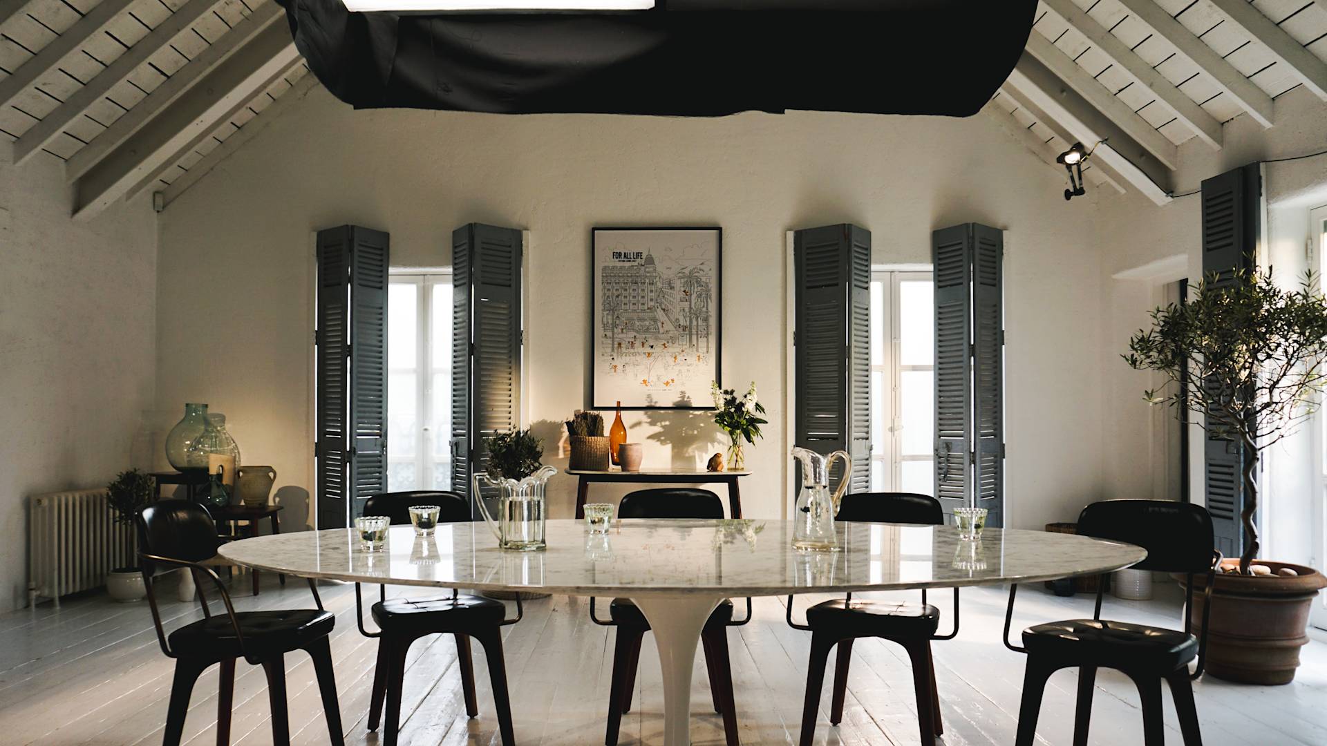 A modern dining room with a large oval marble table, black chairs, glassware, and plants. The space features high ceilings, gray shutters on windows, and a framed artwork on the back wall.