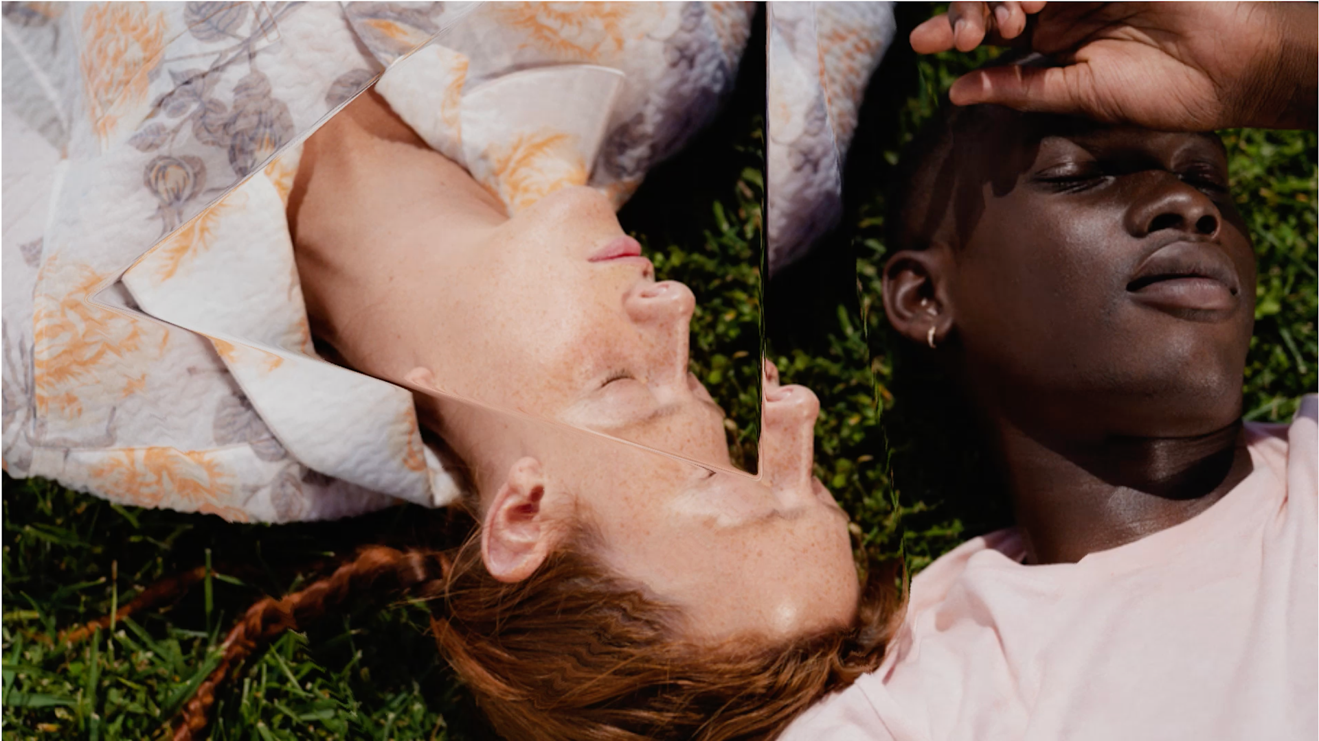 Two people lie on grass, heads close together. One holds a mirror over their face, reflecting the other’s face. The image features contrasting skin tones and soft natural light, creating a dreamy, artistic effect.