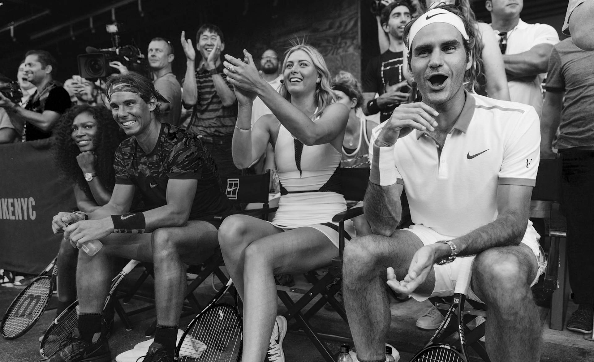 A group of professional tennis players sits courtside, smiling and cheering among an enthusiastic crowd. Some hold rackets and wear tennis attire. The scene captures excitement and camaraderie at a tennis event.