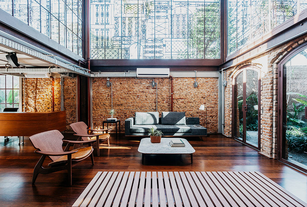A spacious, modern living room features exposed brick walls, large windows with decorative metalwork, wooden floors, a grey sofa, two wooden chairs, a marble coffee table, and abundant natural light.