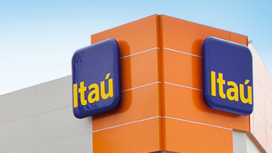 The image shows the corner of a building with two blue signs displaying the word Itaú in yellow letters, set against orange and white walls under a clear blue sky.