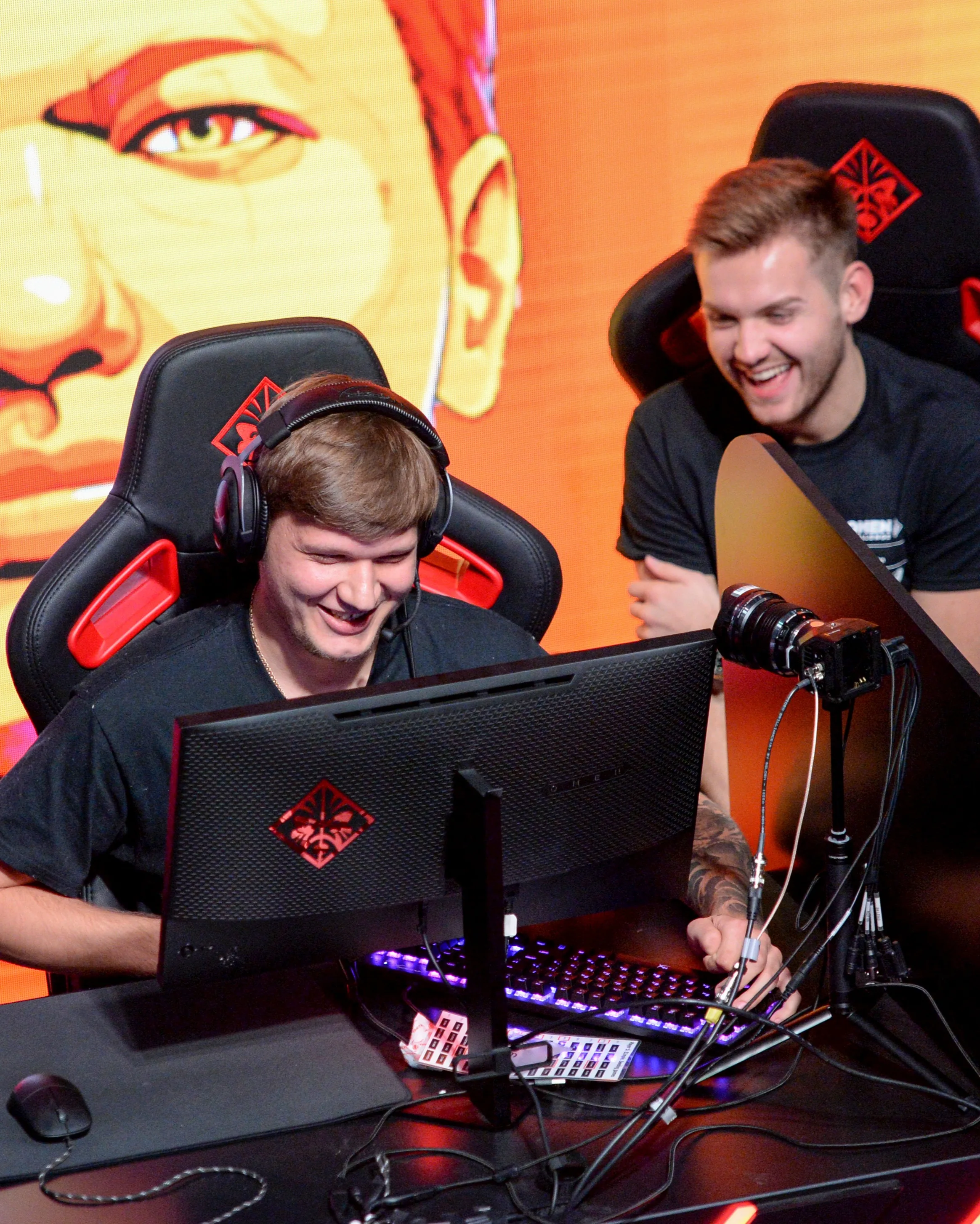 Two young men smiling and laughing while sitting at gaming computers with headsets on, in front of an orange backdrop with a large illustrated face.