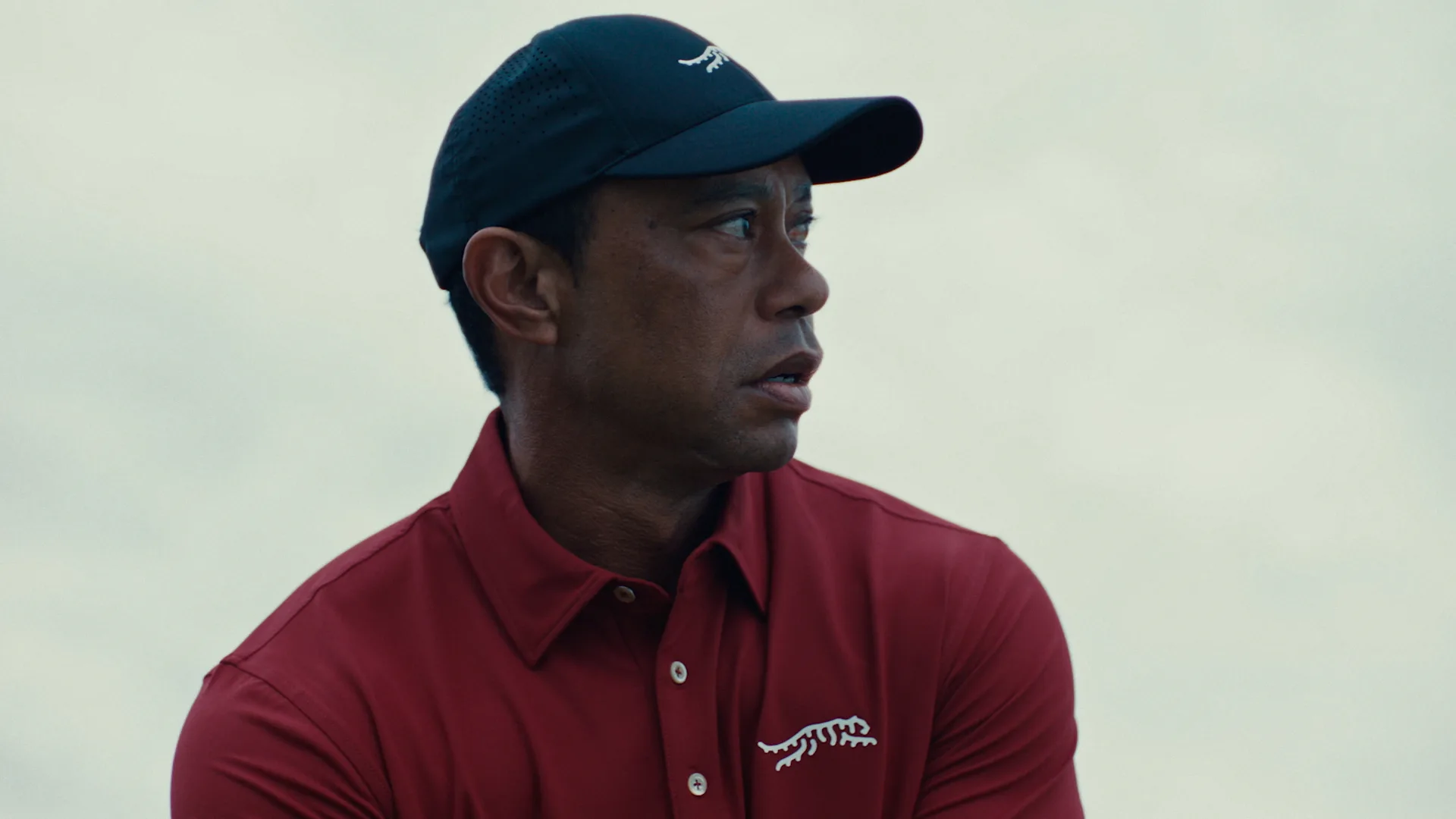 A man in a red collared shirt and dark cap looks to the side with a focused expression. The shirt and cap feature a white animal logo, and the background is softly blurred.