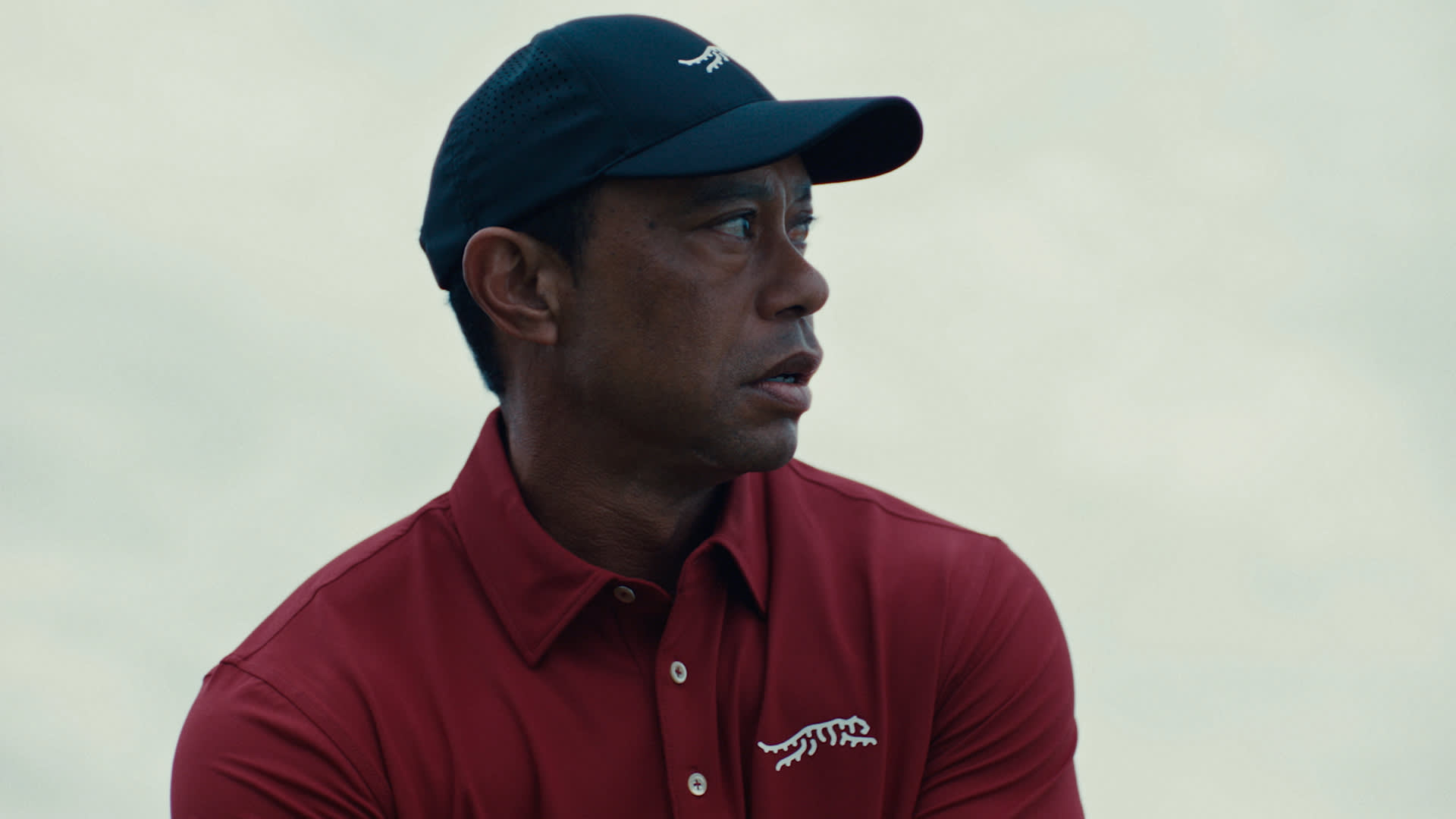 Tiger Woods in a red collared shirt and dark cap looks to the side with a focused expression. The shirt and cap feature a white animal logo, and the background is softly blurred.