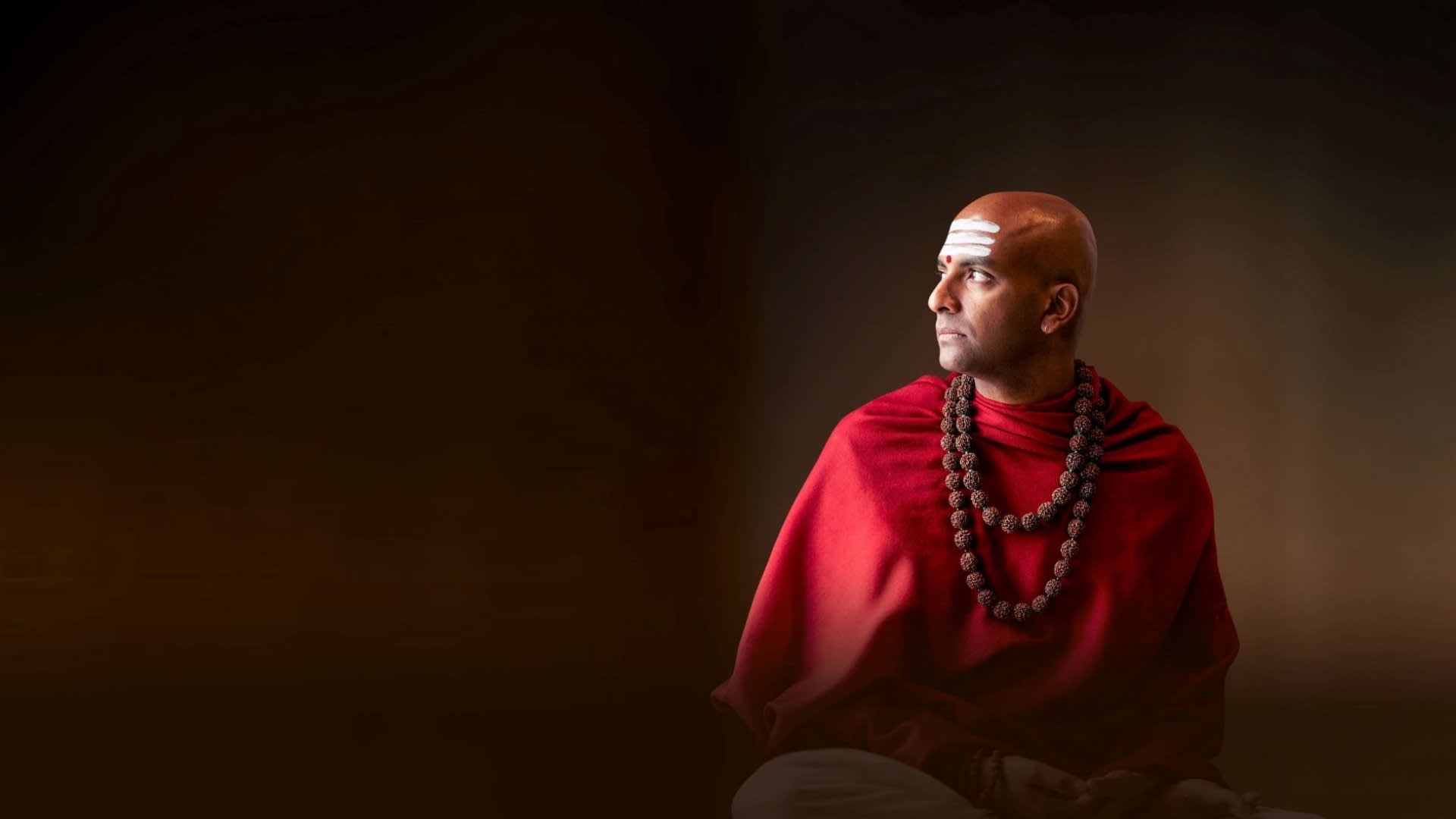 A bald man wearing a red robe and rudraksha necklaces sits gazing to the right with white markings on his forehead, against a dark brown gradient background.
