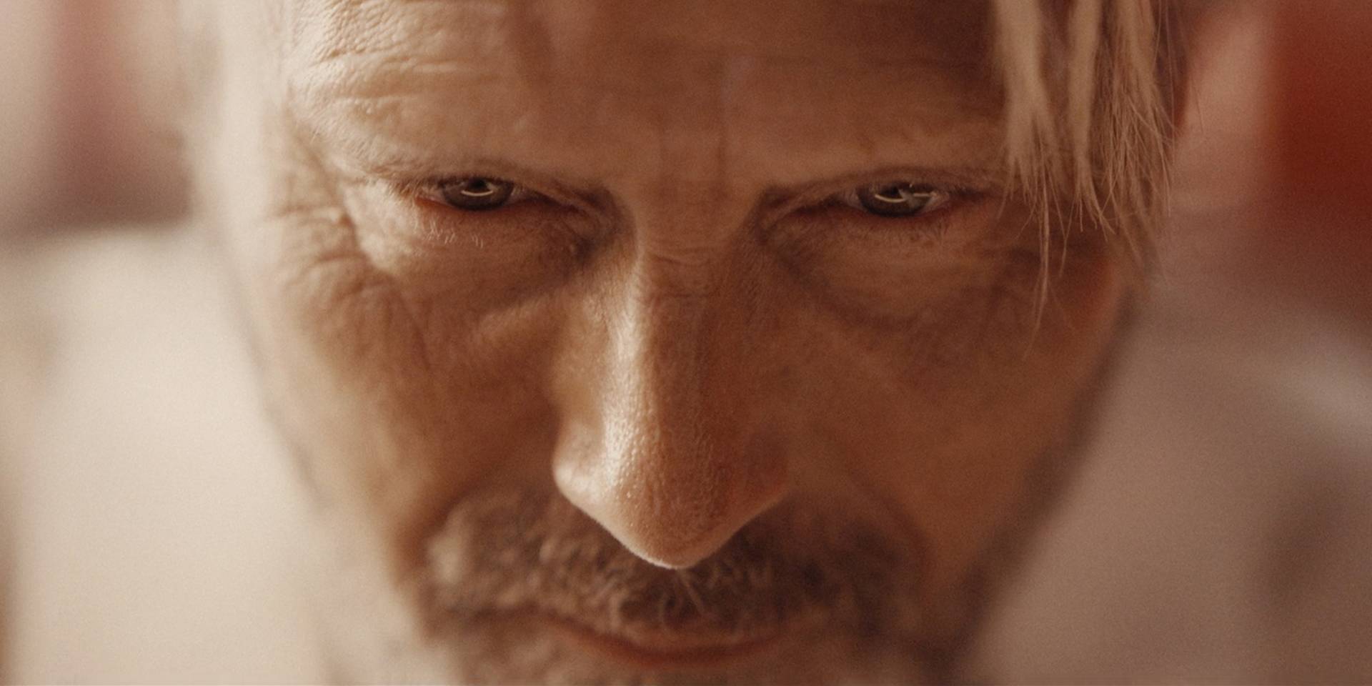 Close-up of an older mans face with intense, focused eyes, furrowed brows, and visible wrinkles. His expression is serious, and his light hair partially falls over his forehead. Warm lighting highlights his features.