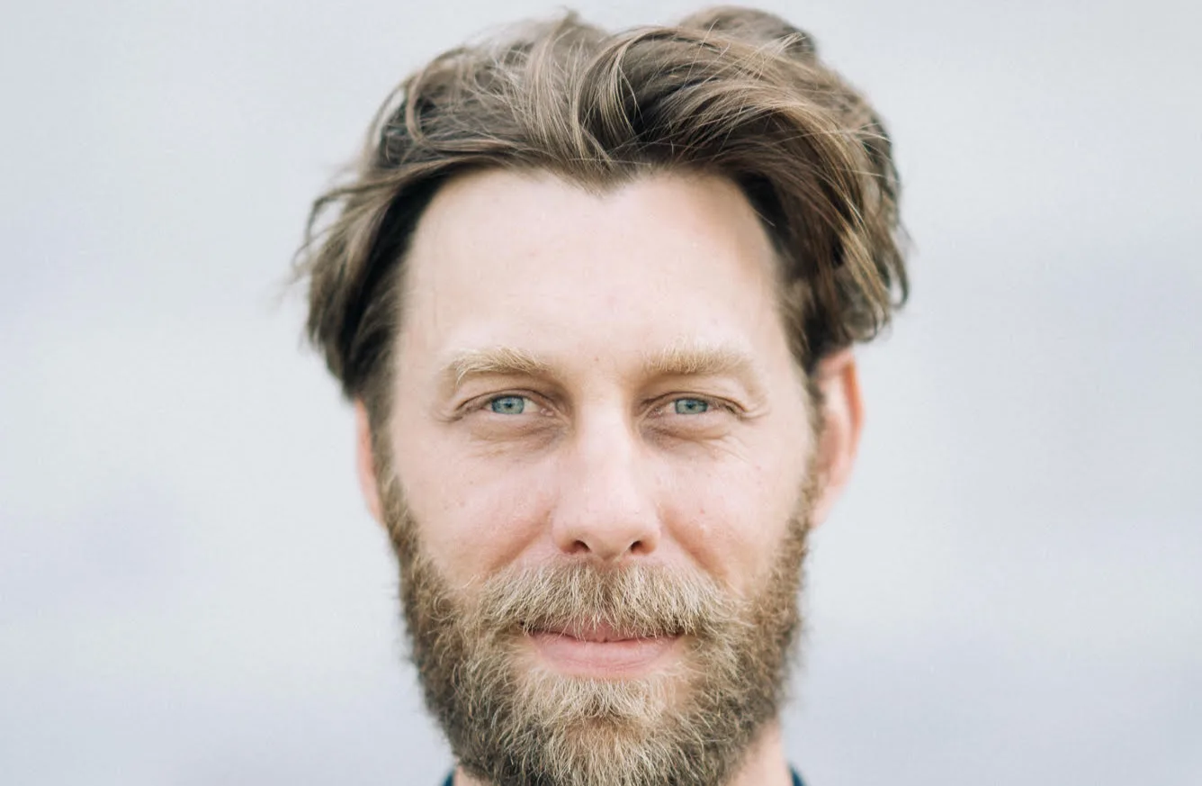 A man with light skin, blue eyes, and brown hair wears a full beard and mustache, looking directly at the camera with a slight smile against a neutral, blurred background.