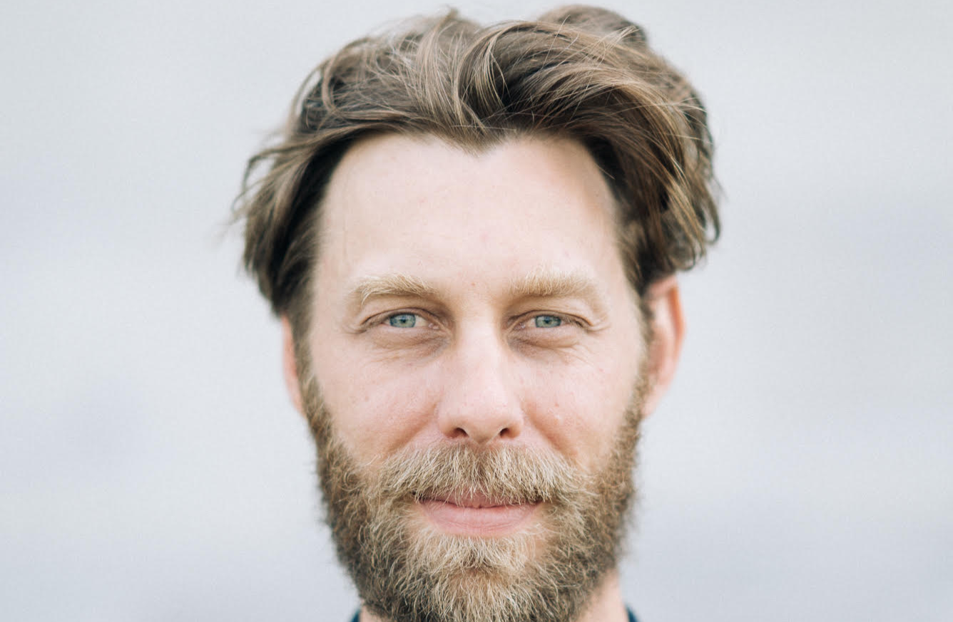 A man with light skin, blue eyes, and brown hair wears a full beard and mustache, looking directly at the camera with a slight smile against a neutral, blurred background.