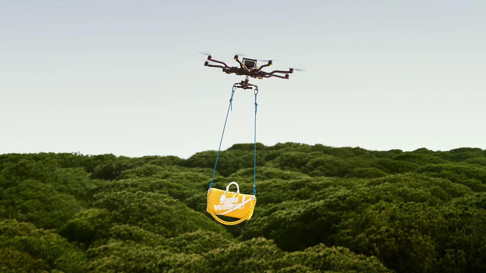 A drone flies over a forest carrying a yellow bag labeled “snacks world” suspended by blue straps. The sky is clear and the trees below are lush and green.