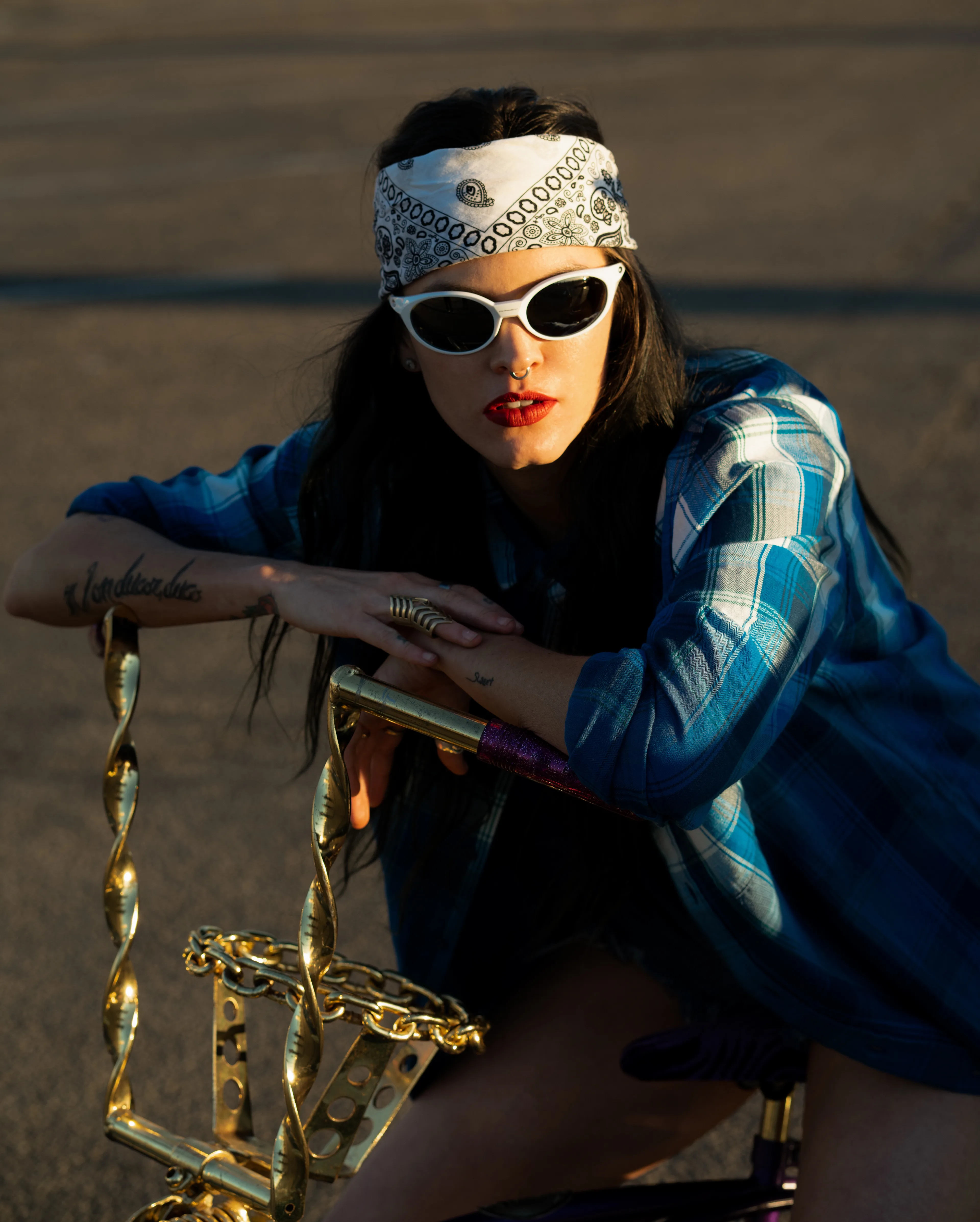 A woman wearing a blue plaid shirt, white sunglasses, and a white bandana leans on the gold handlebars of a bicycle. She has long dark hair, red lipstick, and a tattoo on her right forearm.
