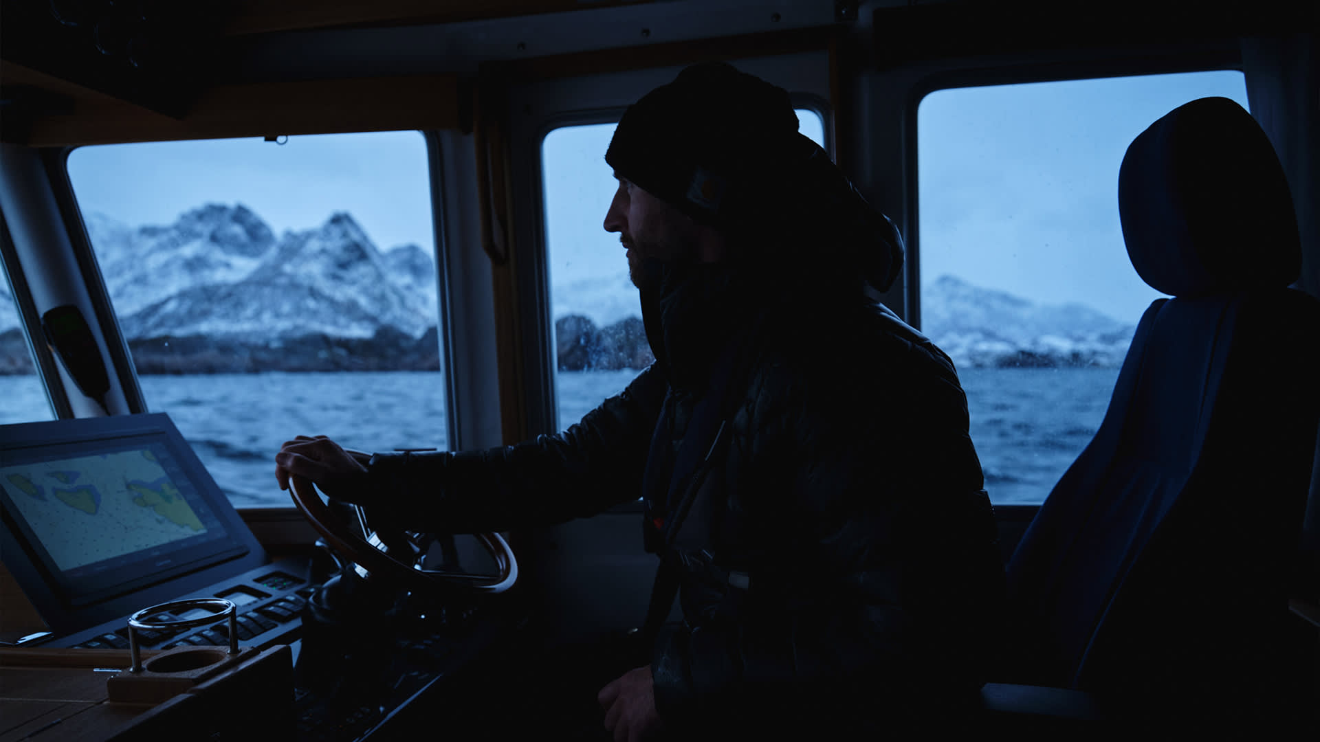 A person wearing a beanie and jacket steers a boat through cold waters, with snow-covered mountains visible through the windows at dusk. The boats control panel displays navigation charts.