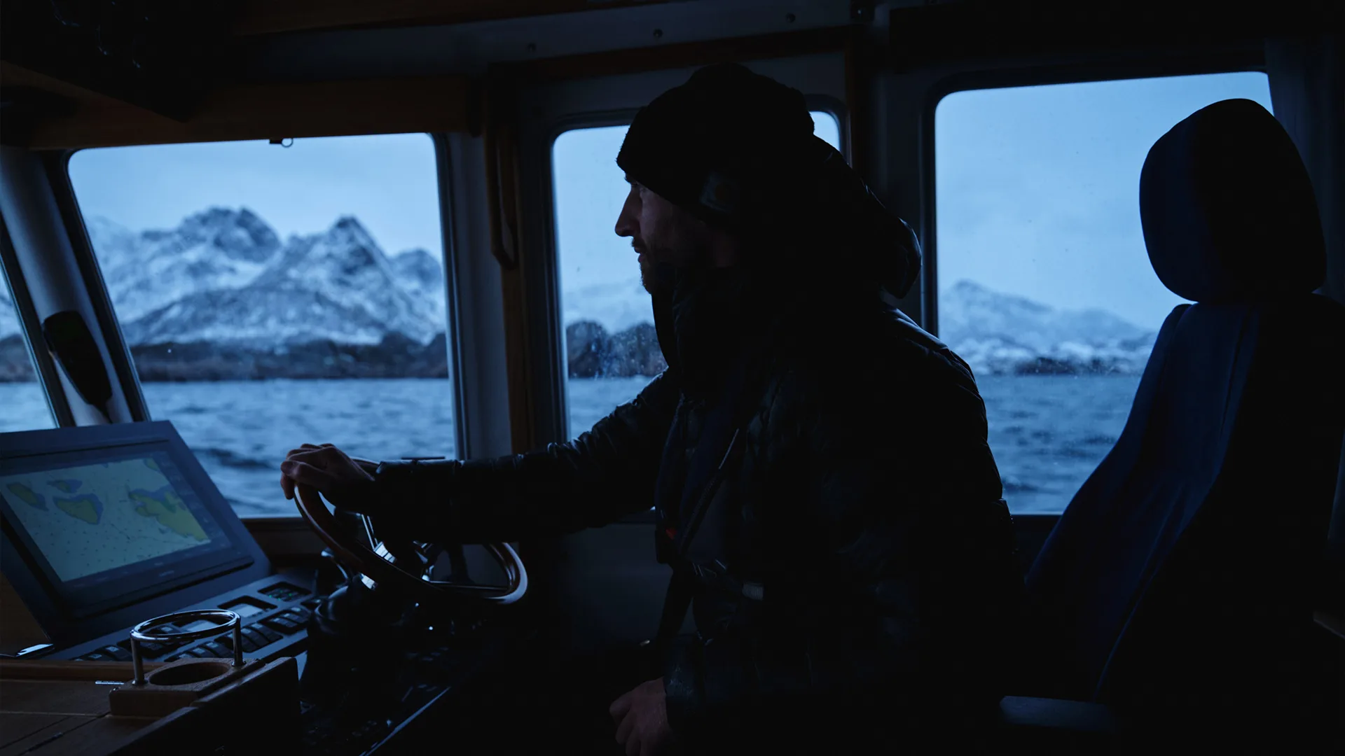 A person wearing a beanie and jacket steers a boat through cold waters, with snow-covered mountains visible through the windows at dusk. The boats control panel displays navigation charts.