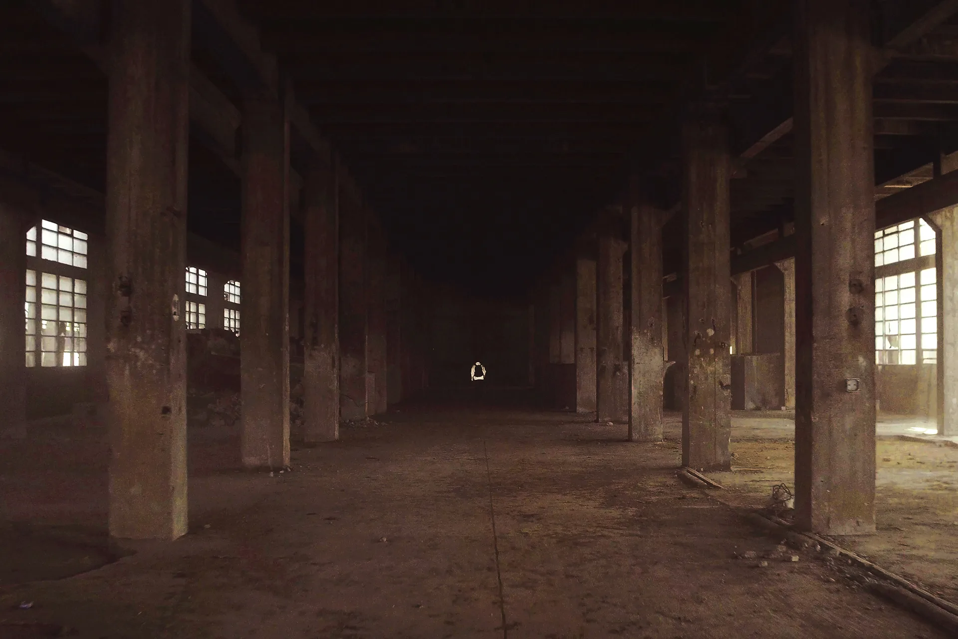 Abandoned, dimly lit industrial building with large concrete pillars, high ceiling, dirty floor, and broken windows. Light filters in from the windows and a small opening at the far end.