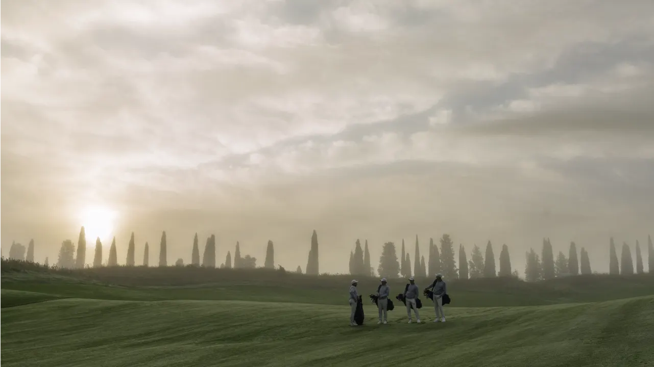 Four people with golf bags stand on a grassy course under a cloudy sky at sunrise or sunset, with tall trees lining the horizon in the background.