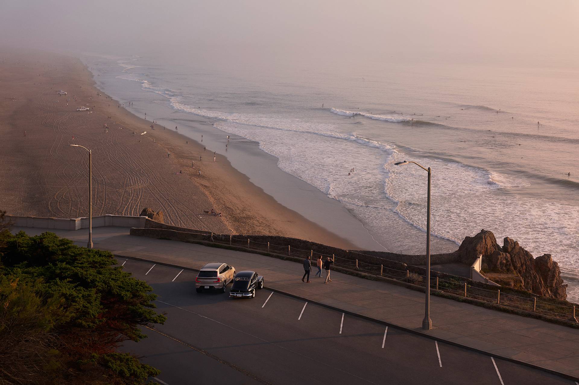 A coastal road with two parked cars overlooks a sandy beach and ocean waves at sunset.