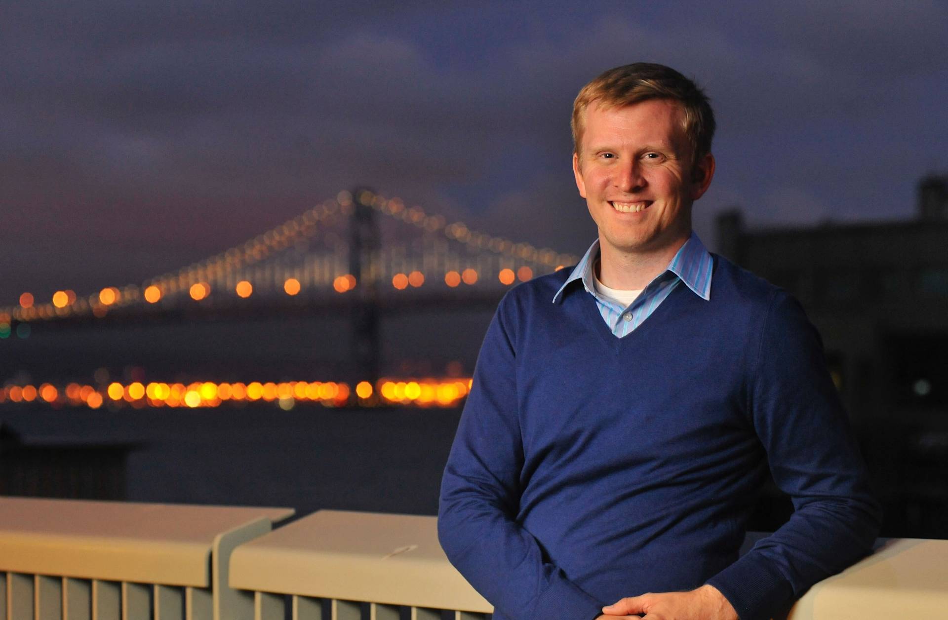 A smiling man in a blue sweater stands in front of a railing at night, with a brightly lit suspension bridge and city lights blurred in the background.