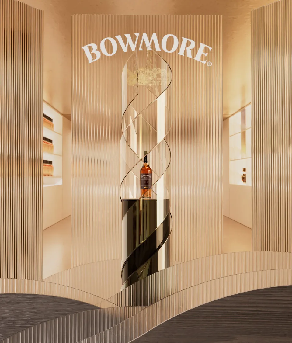 A bottle of Bowmore whisky is displayed in a modern, elegant glass case with a spiral design, set against a sleek, gold and white interior with shelves of bottles in the background. The Bowmore logo is above the display.